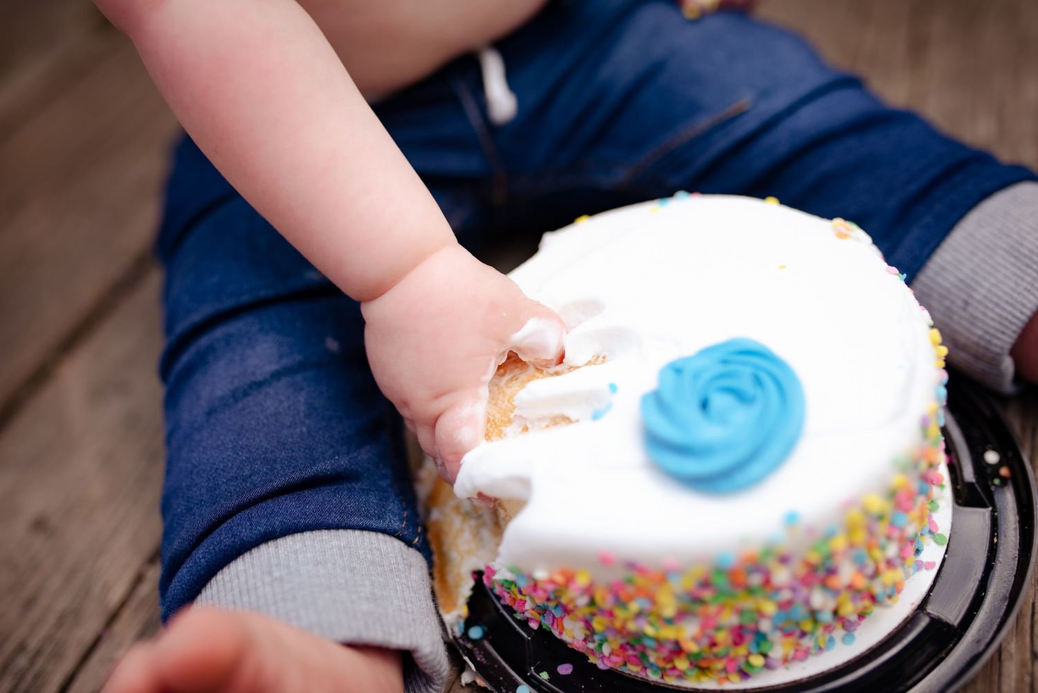 Small hands reach for white frosted birthday cake with blue decoration and sprinkles.