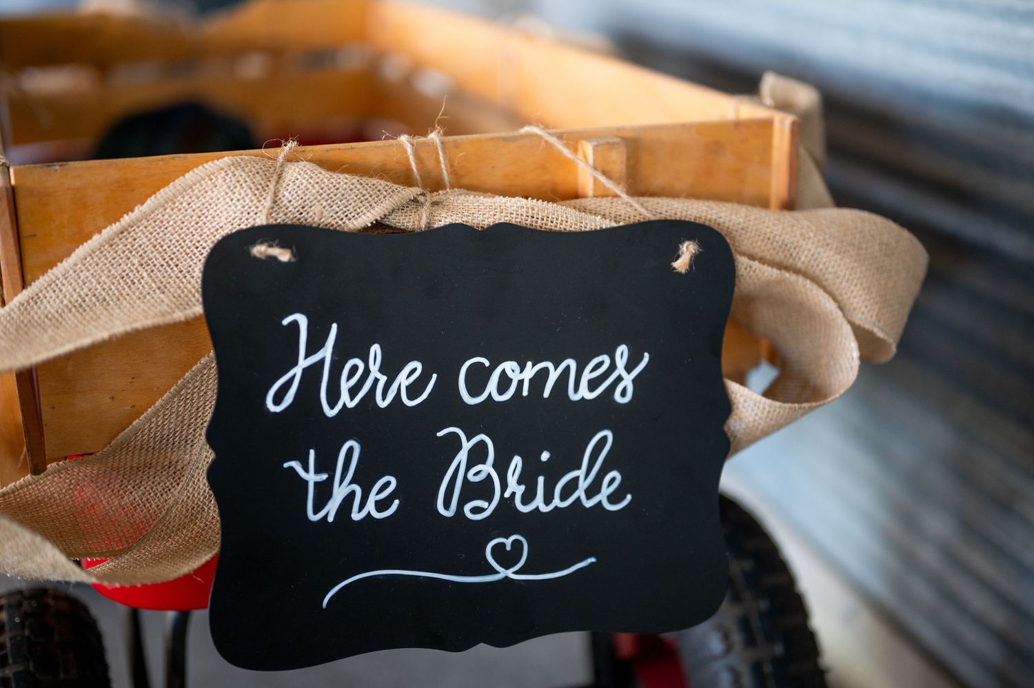 Black chalkboard sign with white handwritten text reading 'Here comes the Bride' with heart decoration.