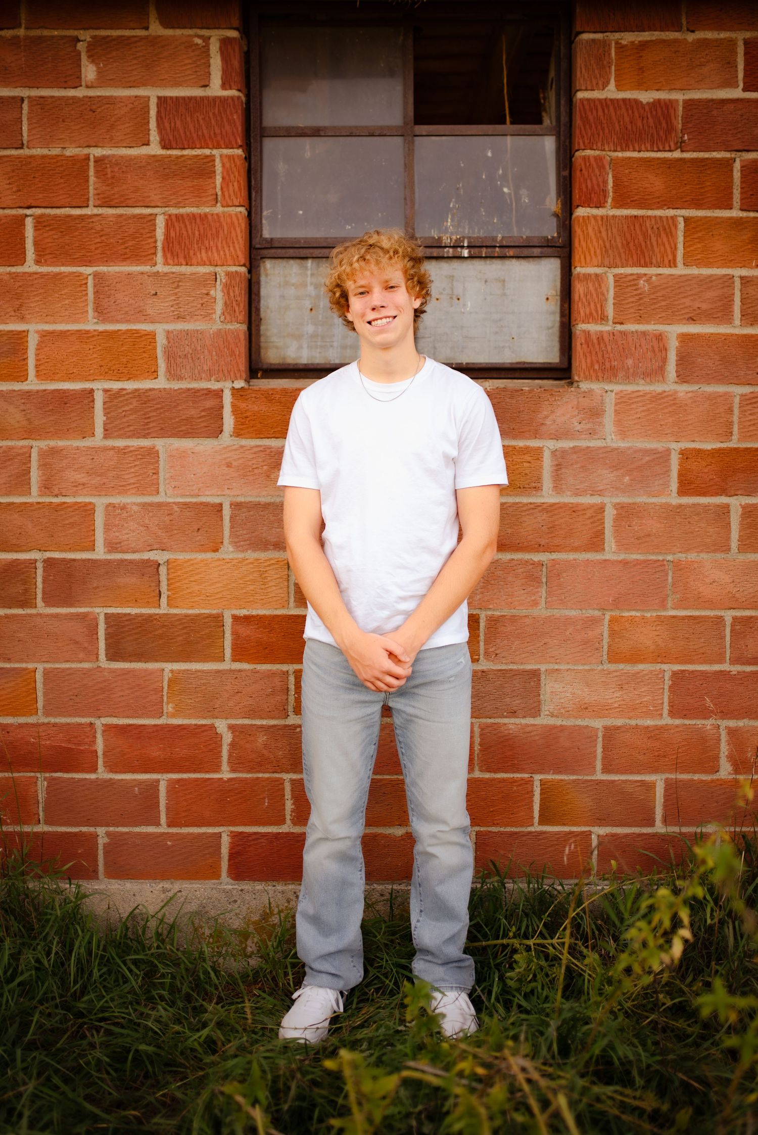 Outdoor portrait against red brick wall with window, wearing white t-shirt and light jeans.