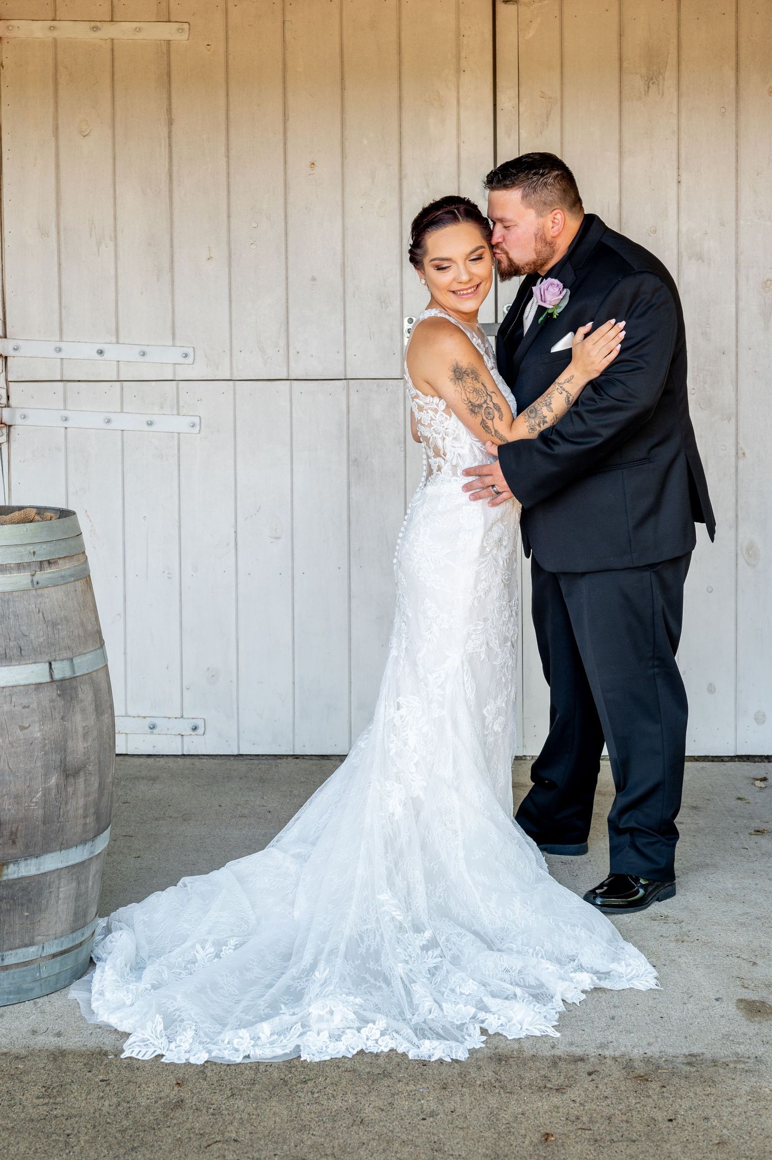 Full length portrait of bride and groom embracing by rustic door with wine barrels.