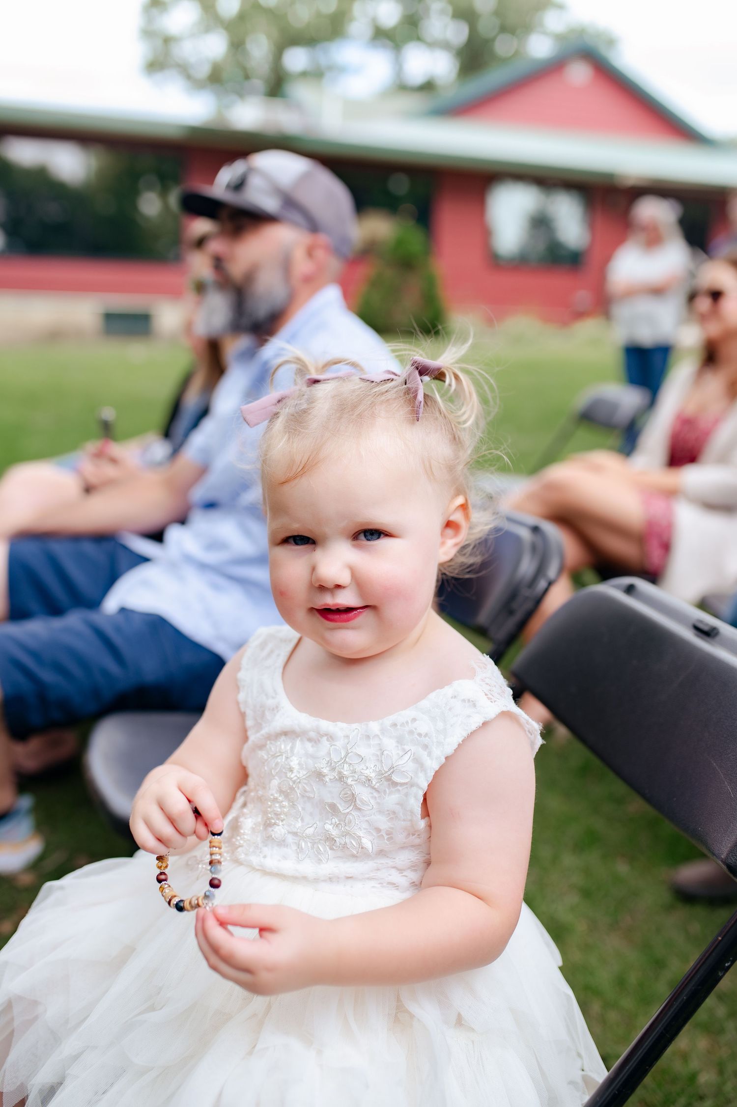 Little flower girl in white tulle dress smiling while sitting at outdoor wedding reception.