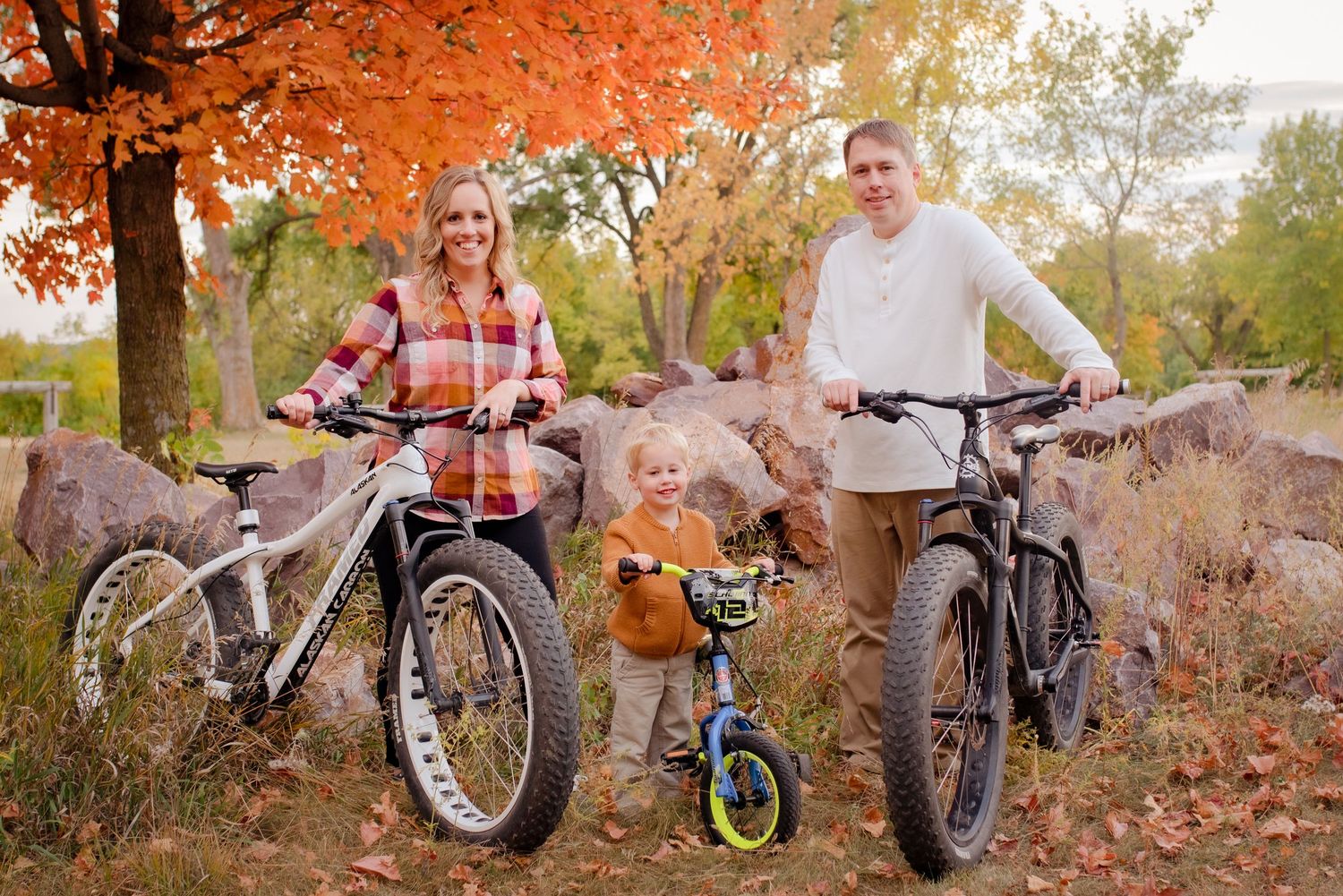 Three people pose with their bikes on an autumn trail surrounded by colorful fall foliage.