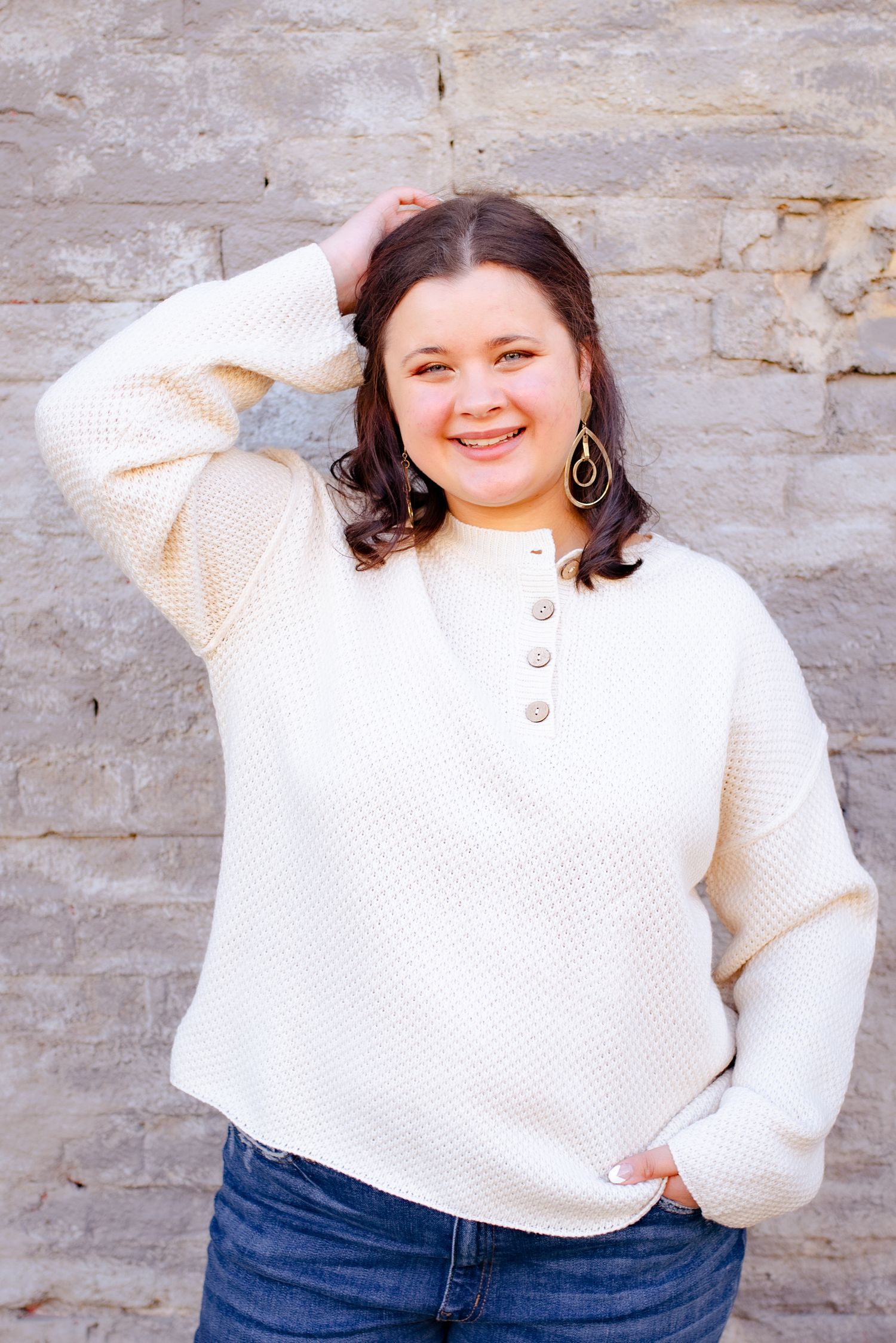 Person in cream knit sweater and dangle earrings poses casually against stone wall.