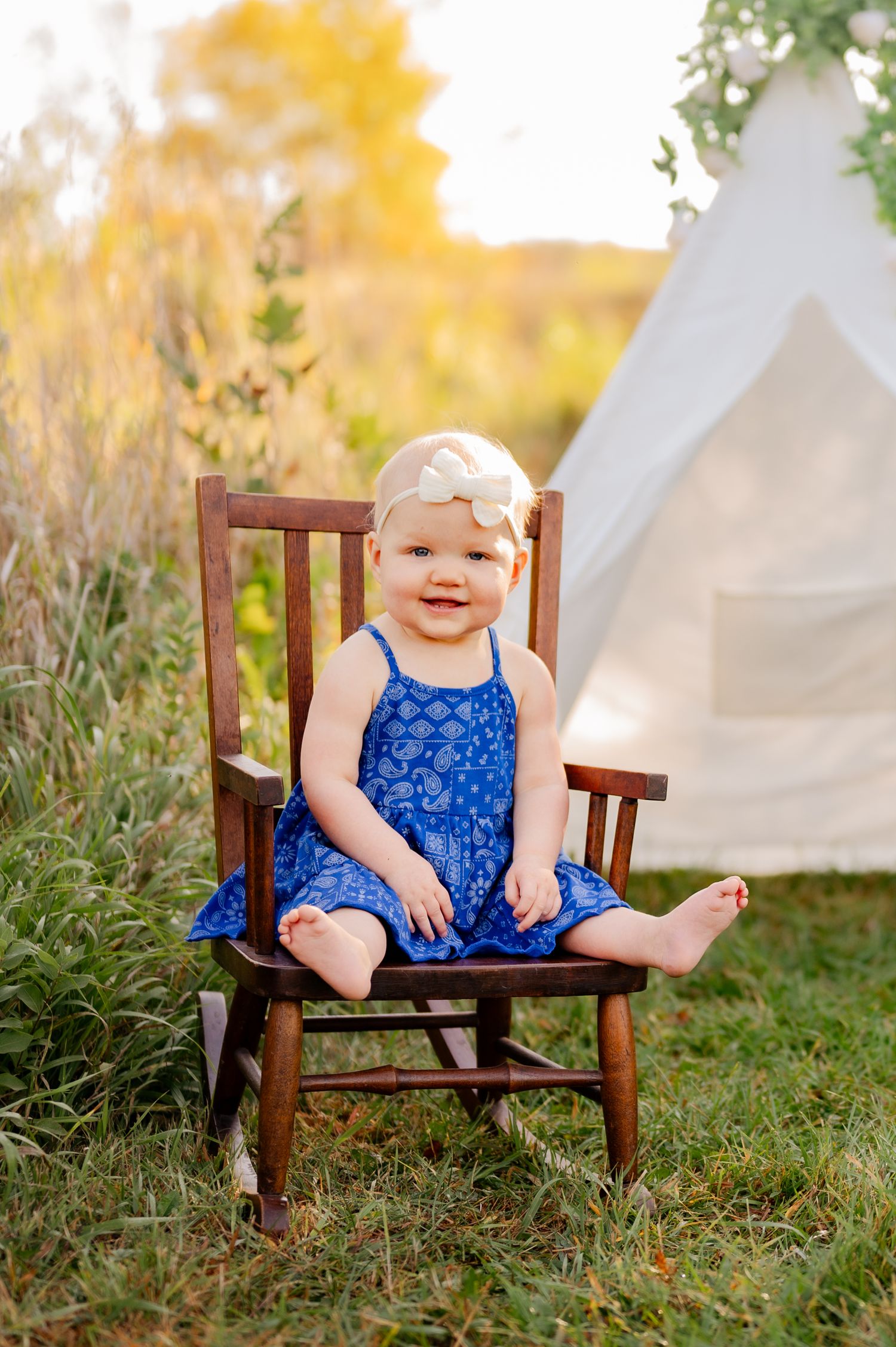 A small child in a navy dress sits in a wooden chair with a white tent backdrop.
