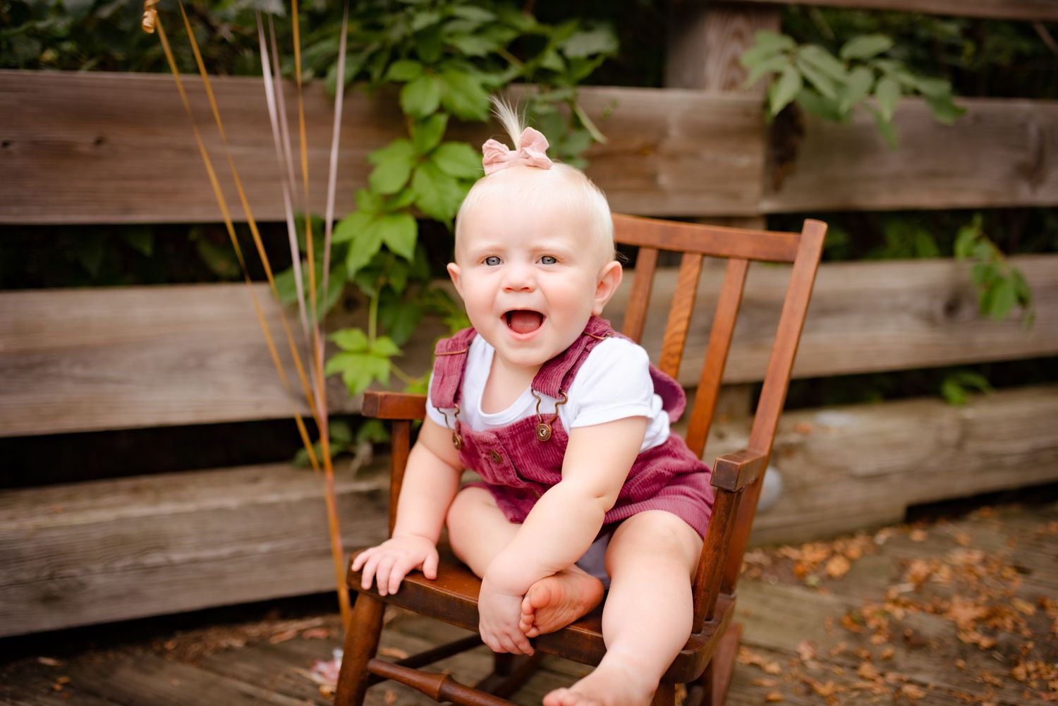 Excited baby wearing burgundy overalls sits in wooden rocking chair outdoors.