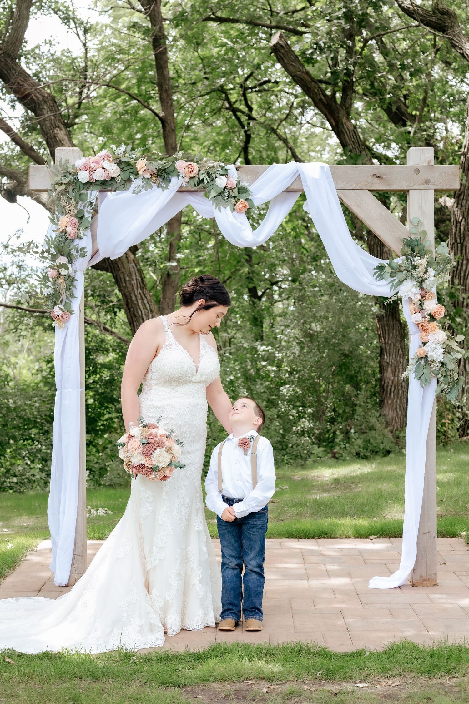 Bride in lace dress stands with young attendant under floral wedding arch.