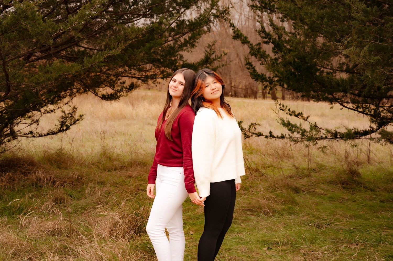 Two people in contrasting red and white tops lean back-to-back in a scenic mountain meadow on a sunny day.