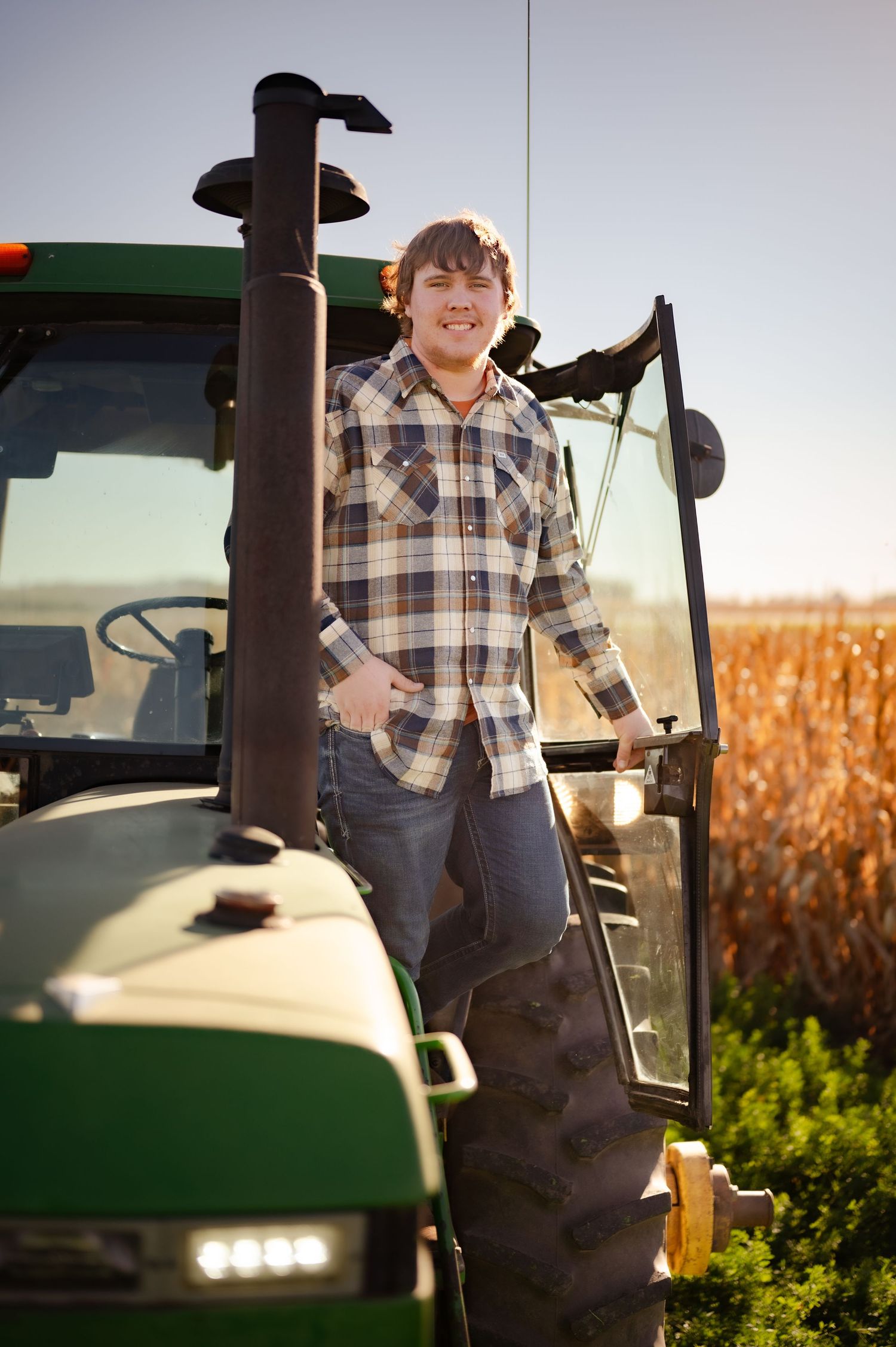 Young child in overalls and plaid shirt stands on a green tractor in a cornfield during golden hour.