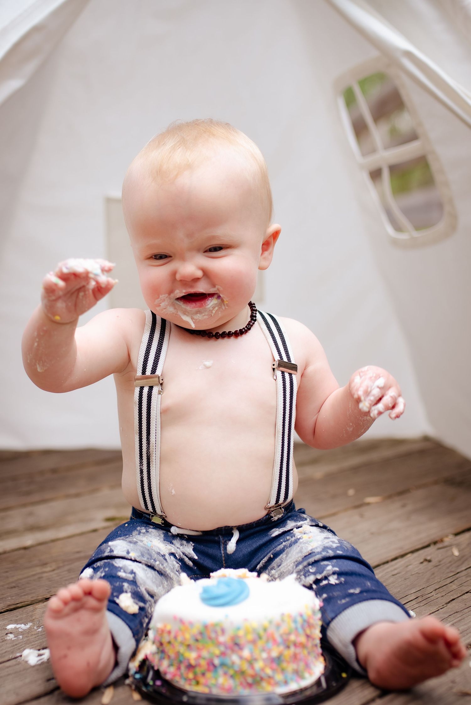 A baby wearing blue shorts and suspenders sits on a wooden floor with a small cake covered in sprinkles.