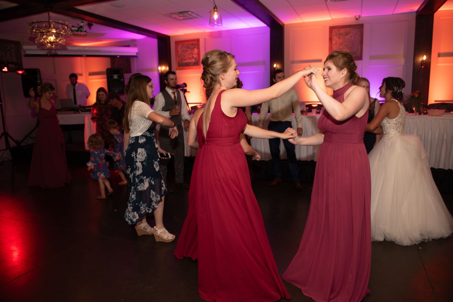 Wedding guests in burgundy bridesmaid dresses dancing together at reception.