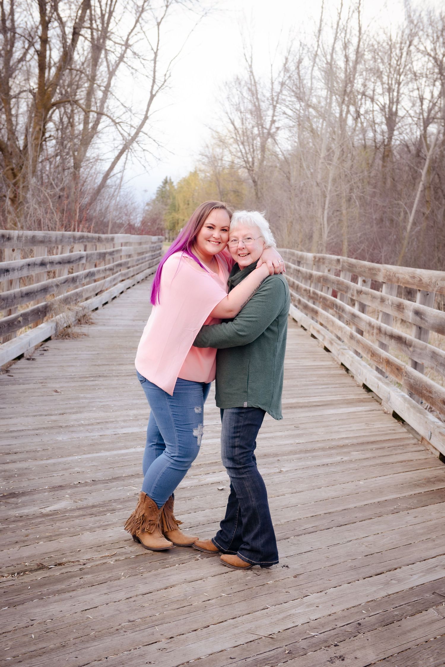 Two people sharing a warm embrace while standing on a wooden boardwalk surrounded by bare trees in winter.