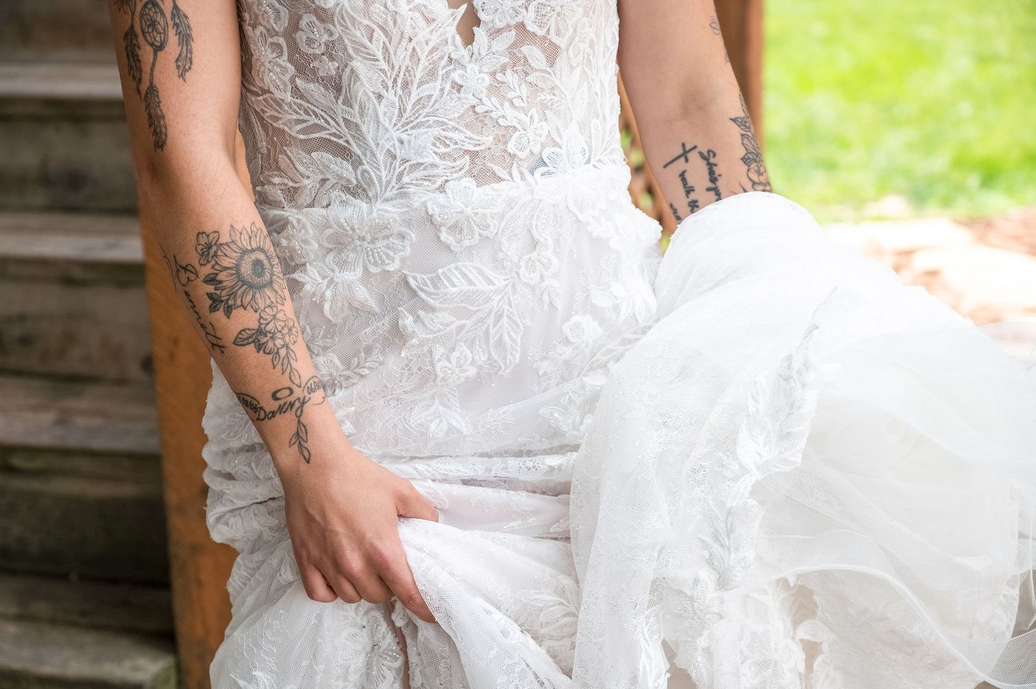 Close-up detail of lace wedding dress and tattooed arms on outdoor stairs.