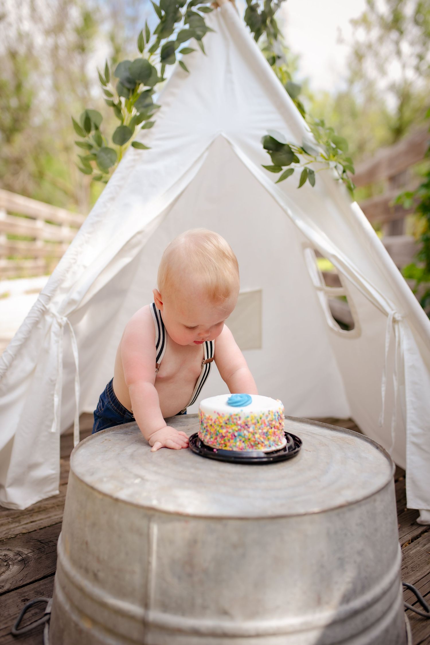 A baby in striped suspenders examines a birthday cake inside a white teepee on wooden deck.