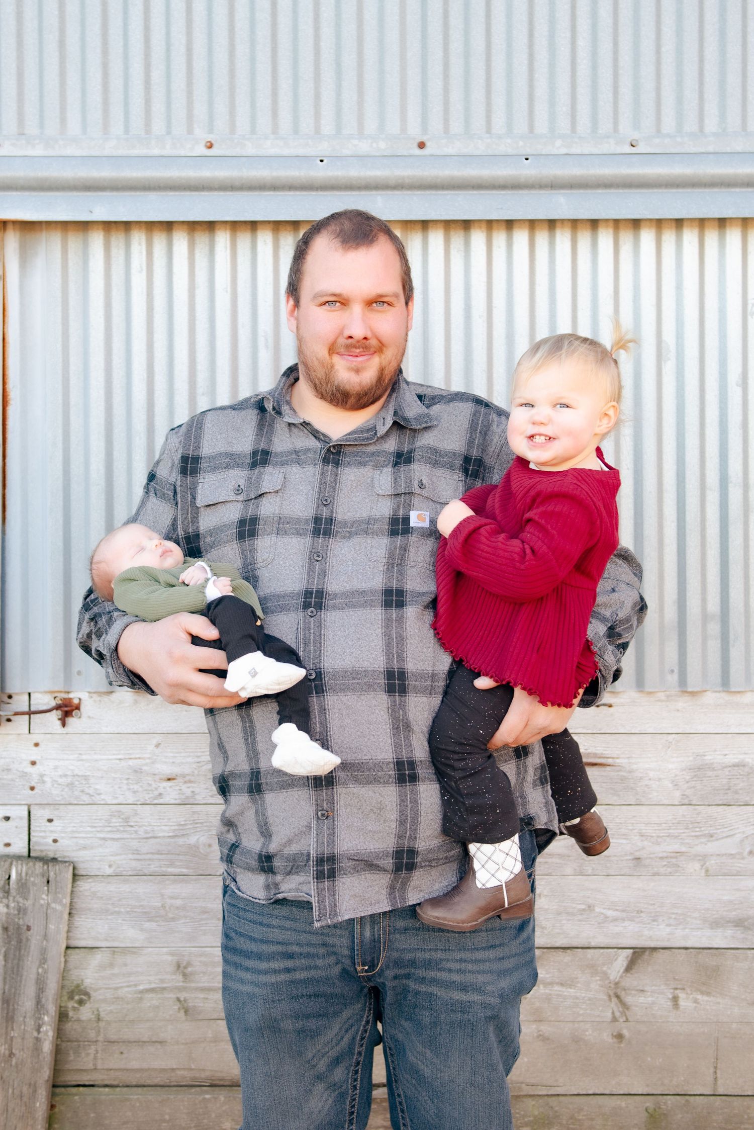 Father in plaid shirt holding two children while standing against corrugated metal background.
