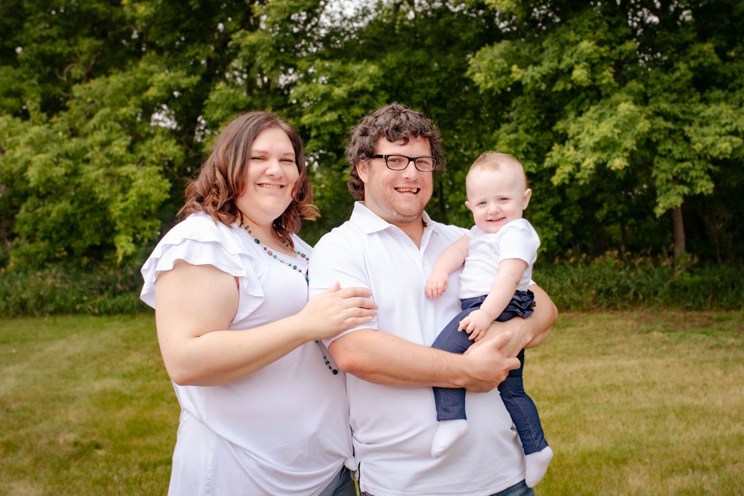 A happy family poses together outdoors with their infant child against a lush green background.