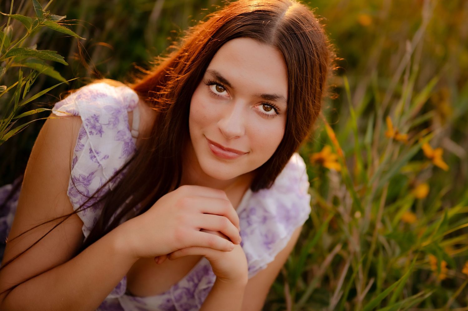 Person with long hair poses in a meadow with warm sunlight filtering through tall grass and flowers.