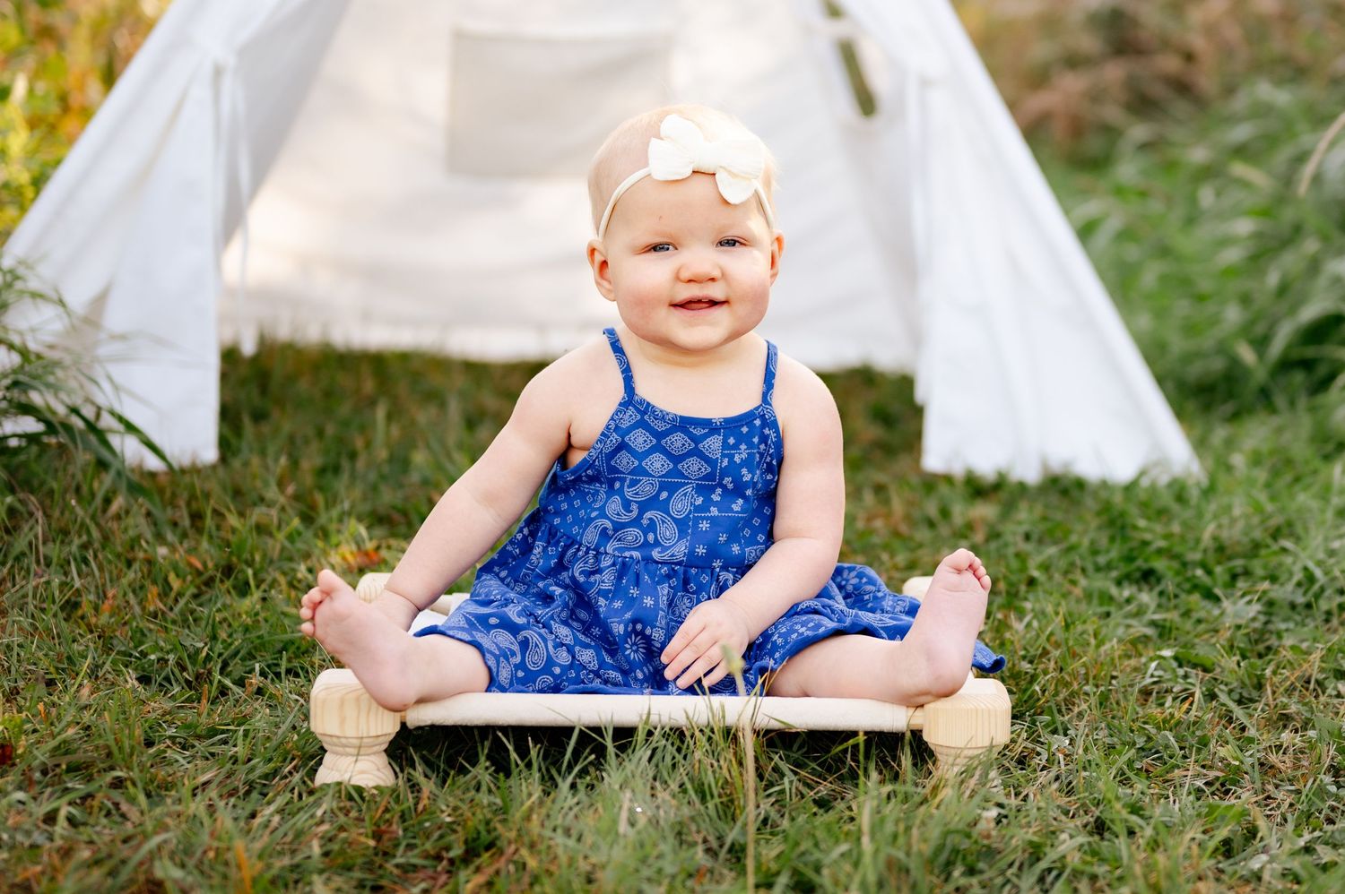 A toddler in a blue dress sits on a white platform with a teepee tent in the background.