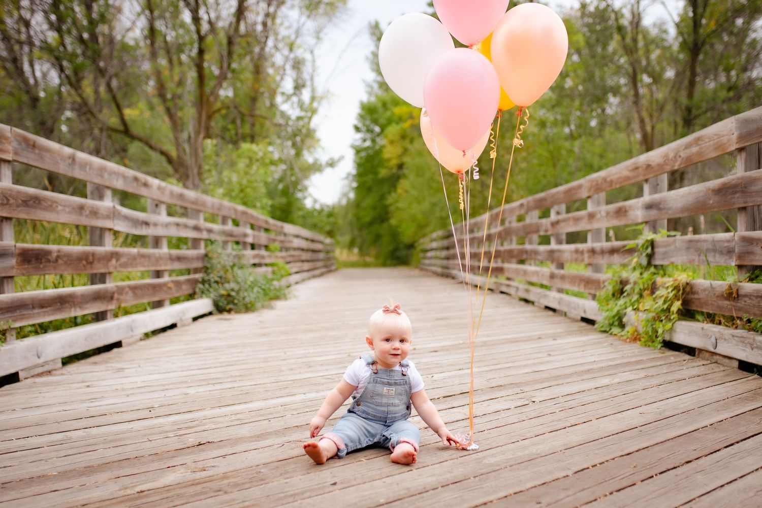 Baby sits on wooden bridge with pink and peach balloons in long perspective shot.