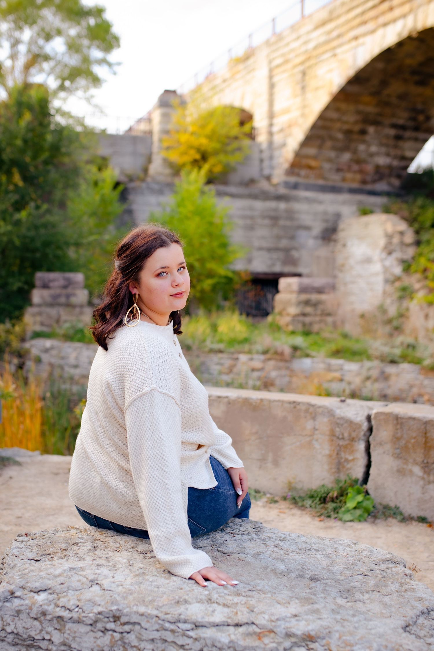 Person in white sweater sits on rock near stone bridge with autumn foliage in background.
