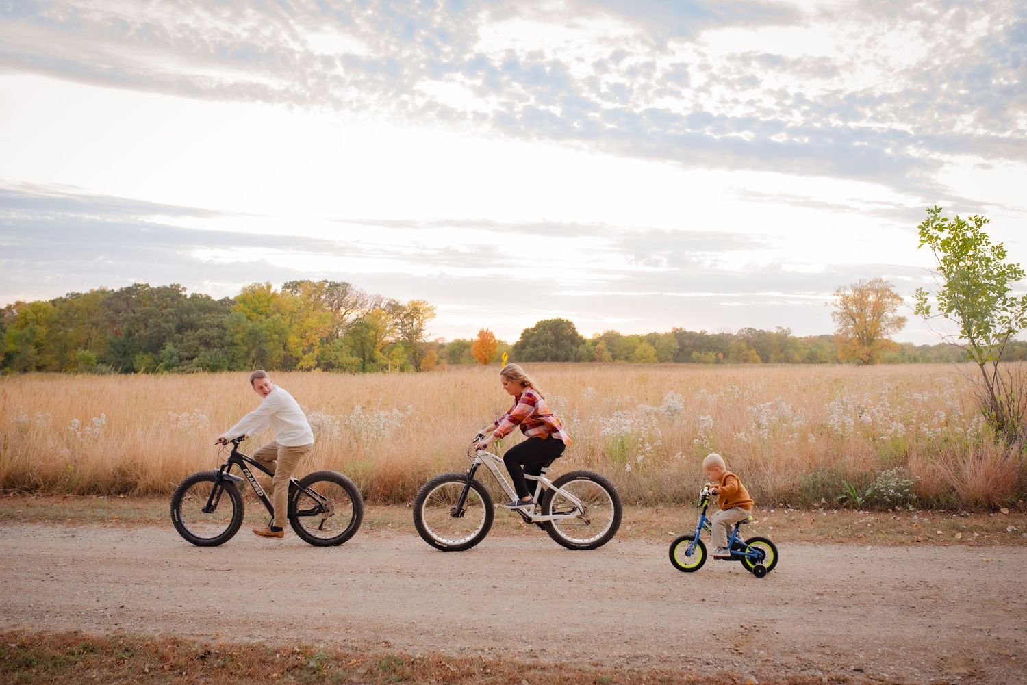 Three cyclists ride along a dirt path during sunset with autumn colors in the background.