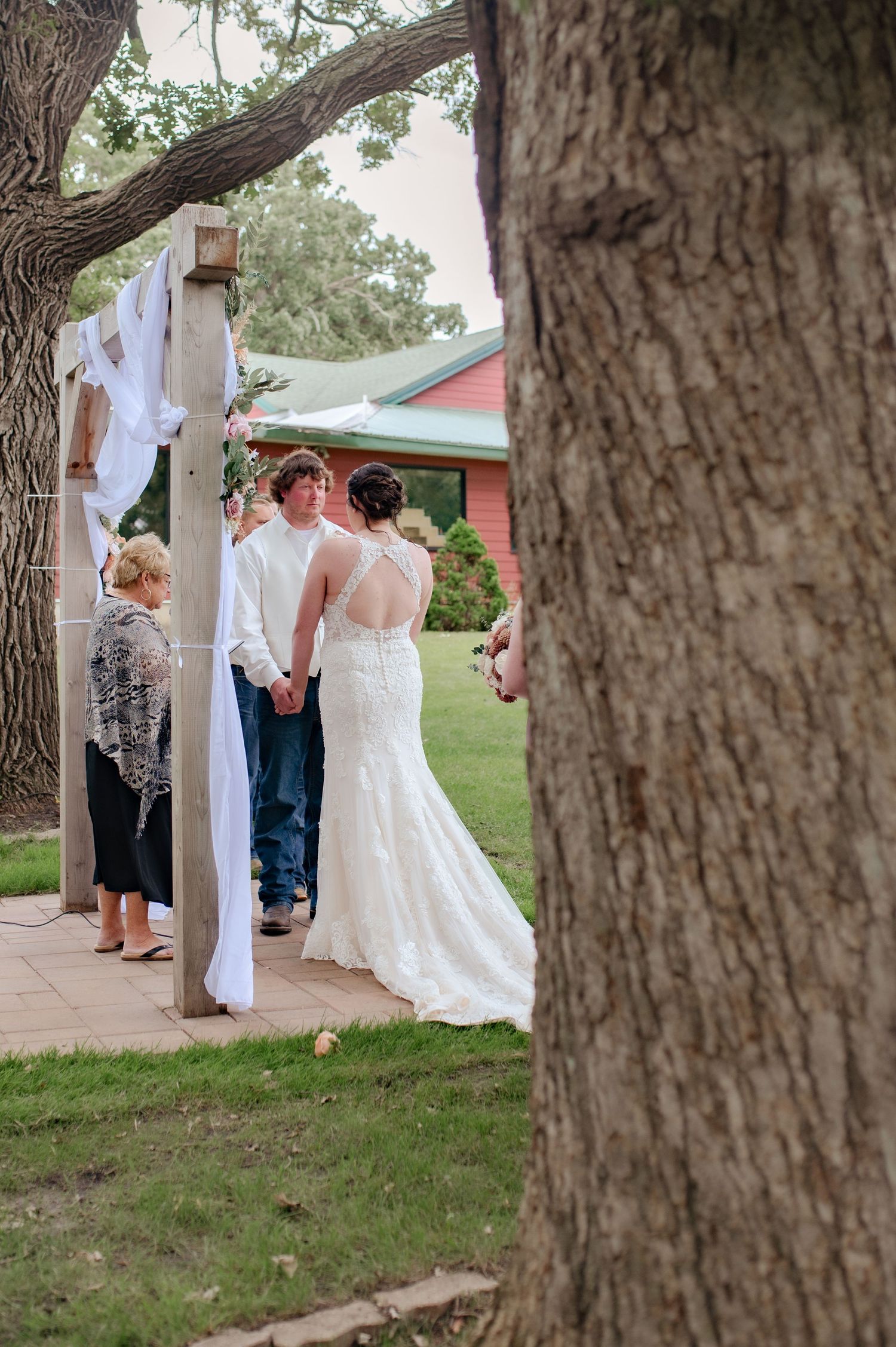 Wedding ceremony glimpse through trees showing decorated arch and guests at outdoor venue.