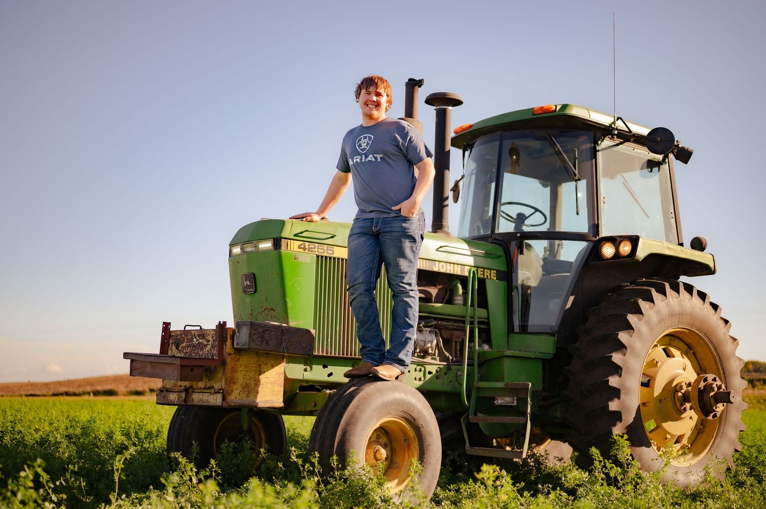 A farmer stands confidently on a green John Deere tractor in an agricultural field during golden hour.