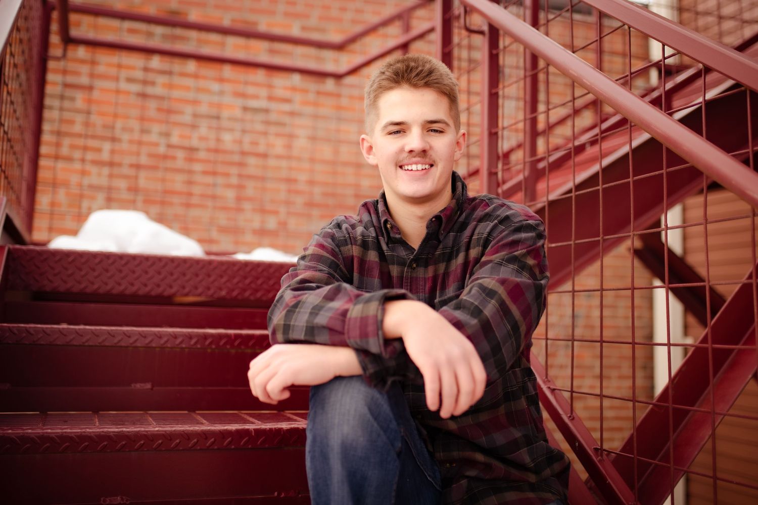 Student in plaid flannel shirt sits casually on red metal stairs.