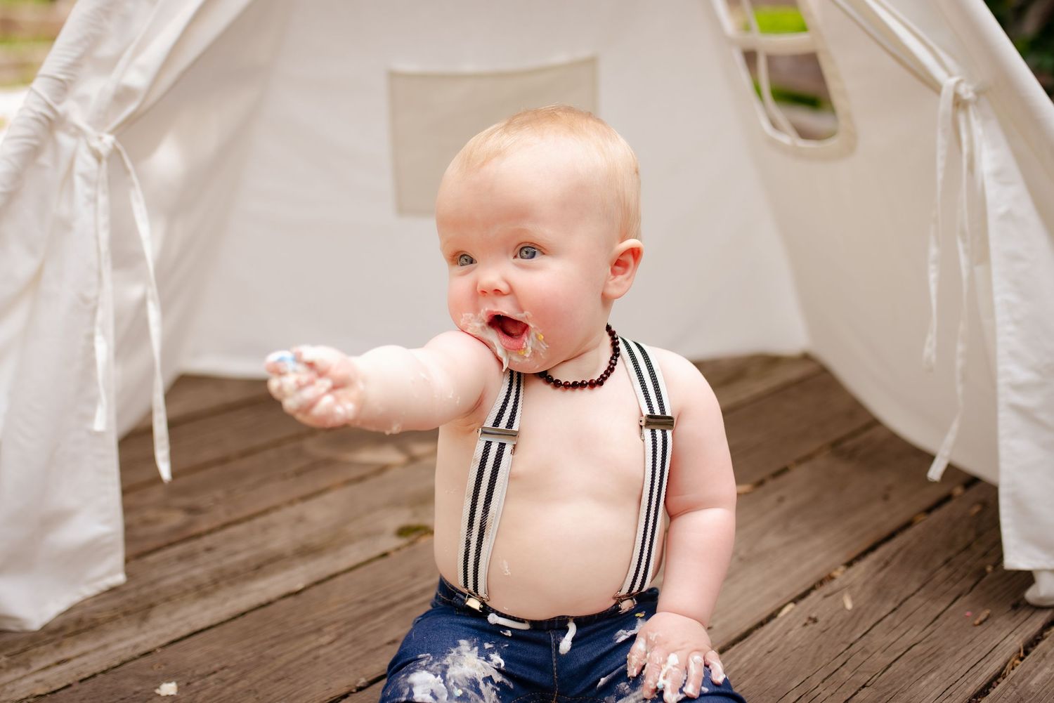 A baby in navy suspenders reaches out while sitting in white teepee tent.