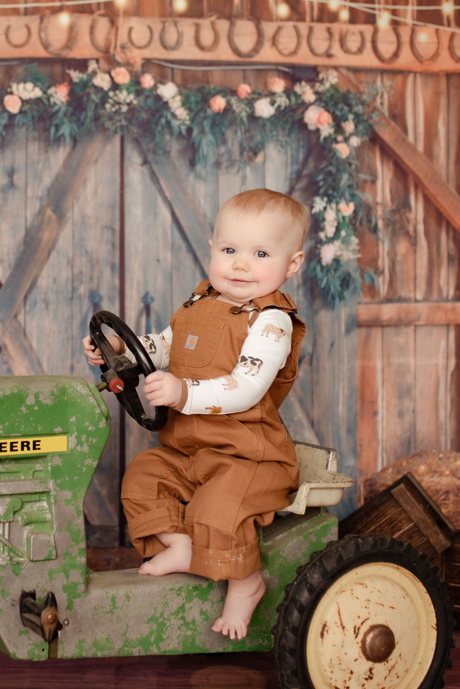 Toddler in brown overalls sits on green John Deere toy tractor in rustic barn setting.