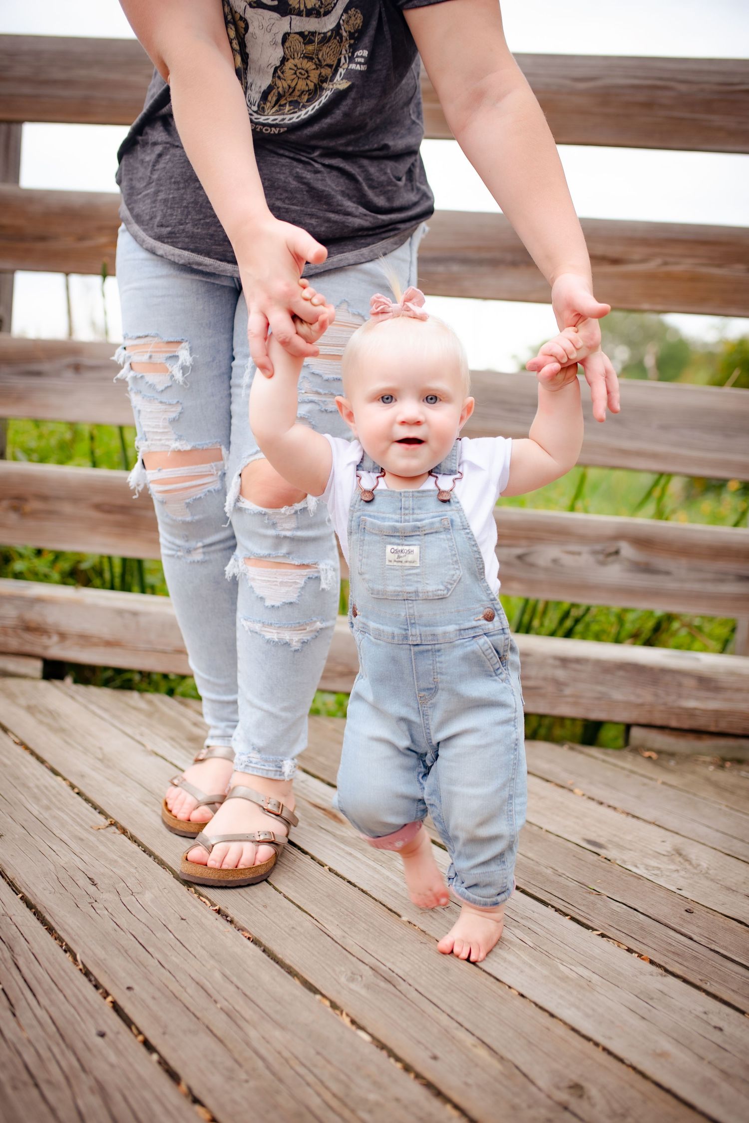 Baby in denim overalls learning to walk while holding parent's hands on wooden deck.