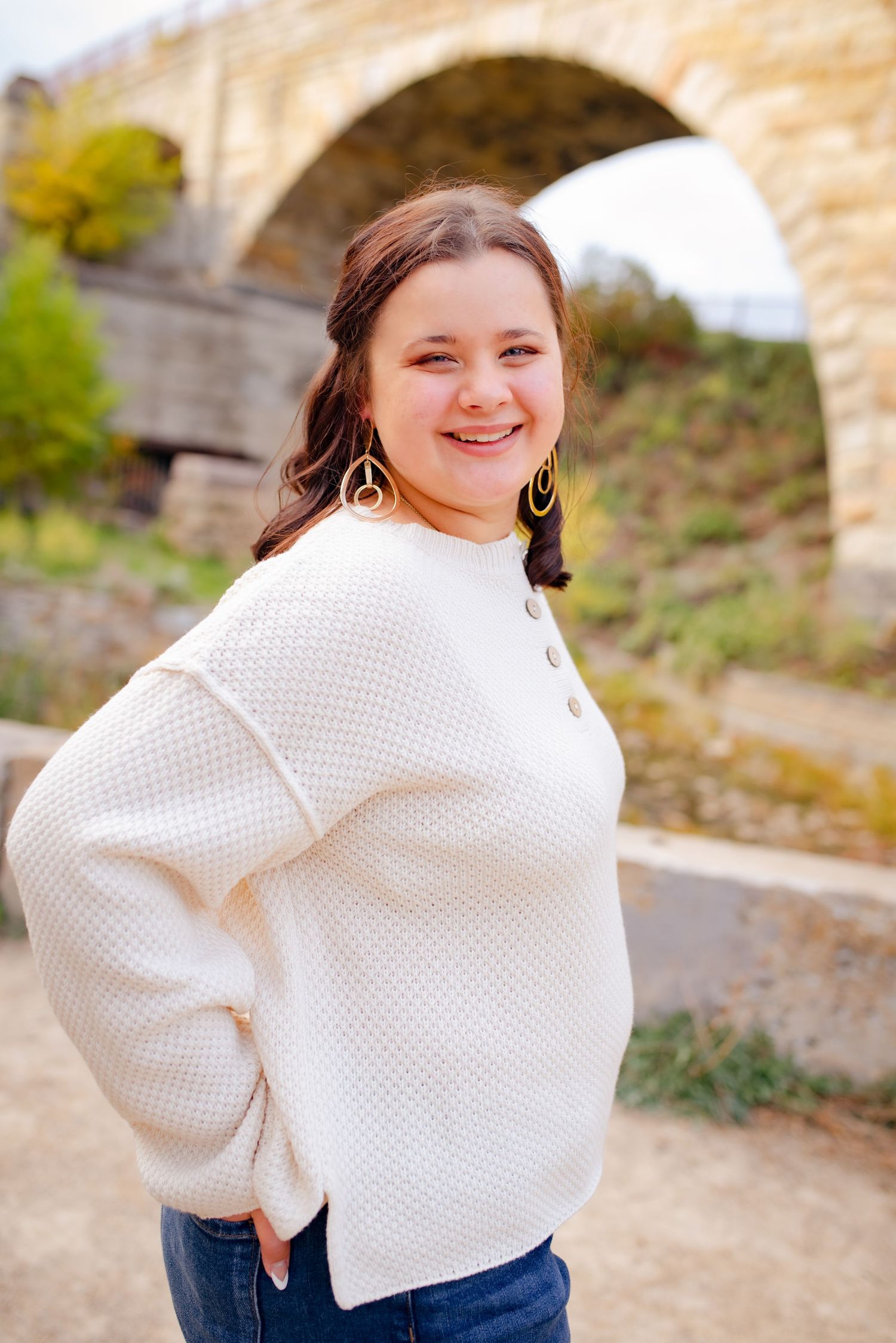 A casual outdoor portrait in front of a stone bridge wearing a white knit sweater and jeans.