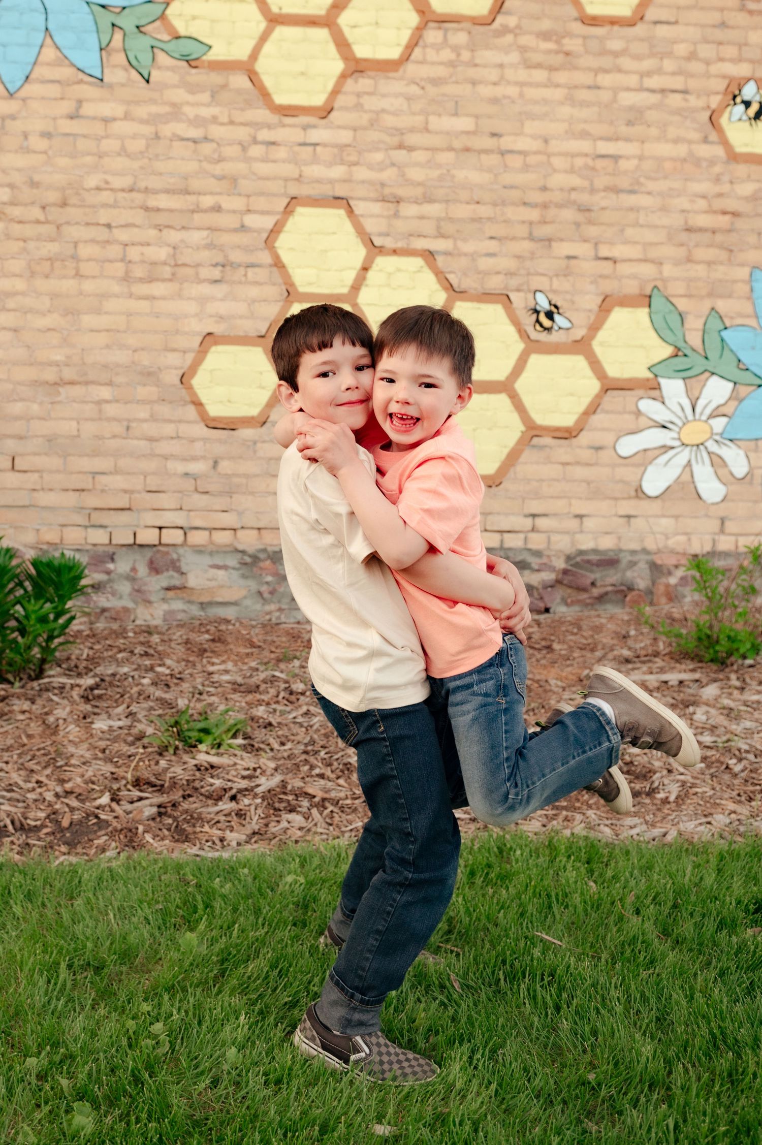 Two children hug and laugh together in front of a decorated brick wall with honeycomb designs.