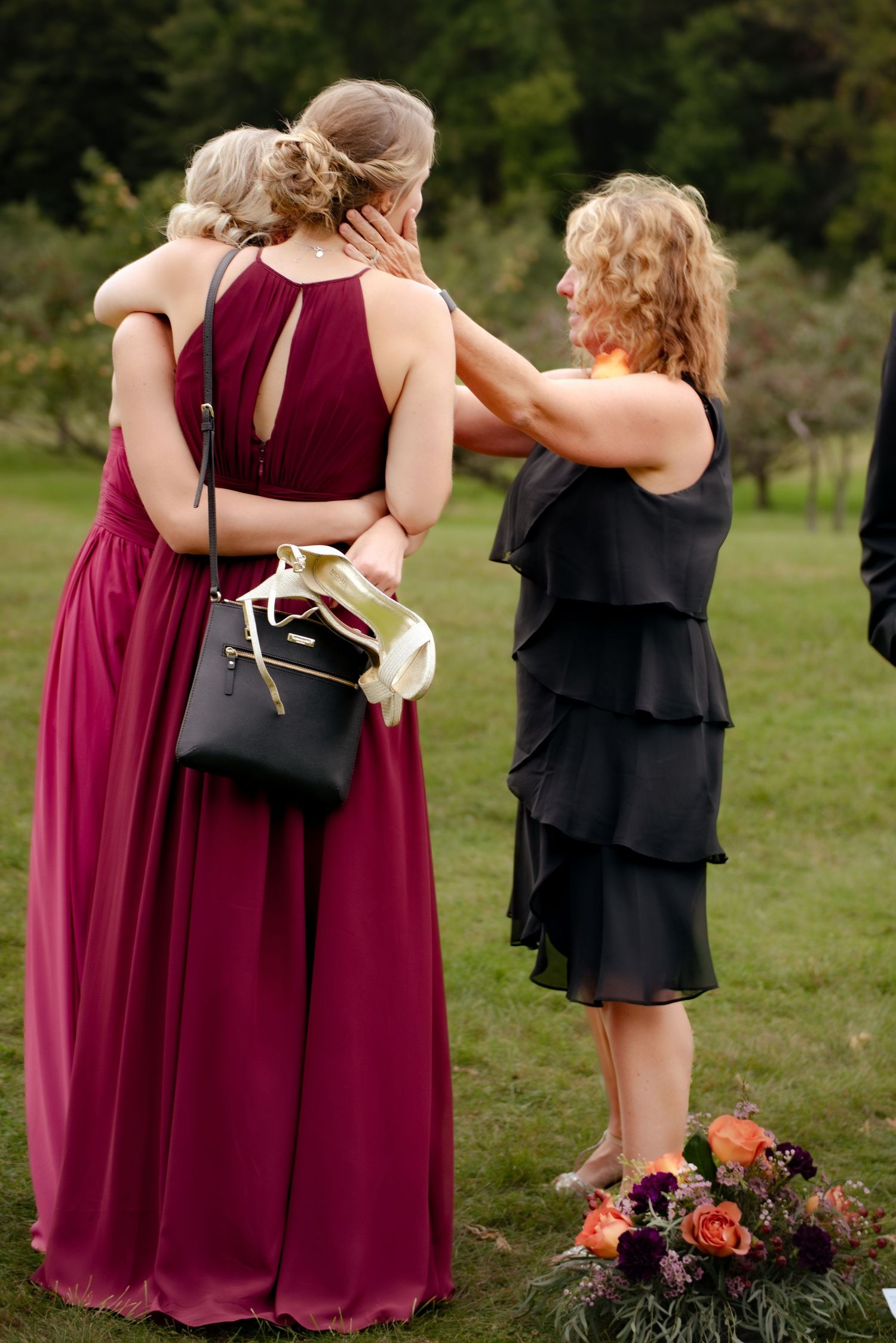 Bridesmaids in burgundy and black dresses gather around a floral arrangement outdoors.
