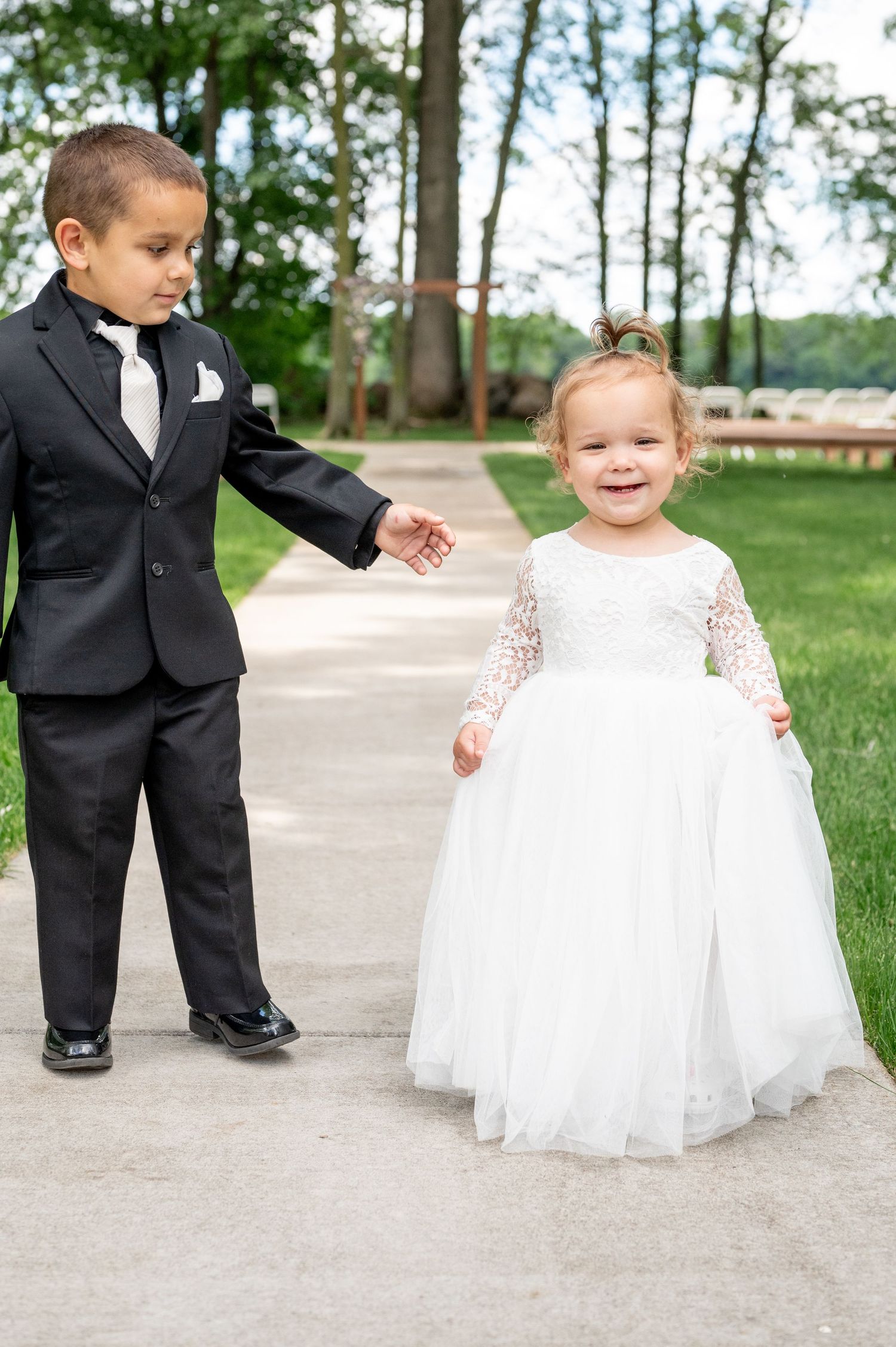 Ring bearer in tuxedo and flower girl in white dress walk down outdoor path.