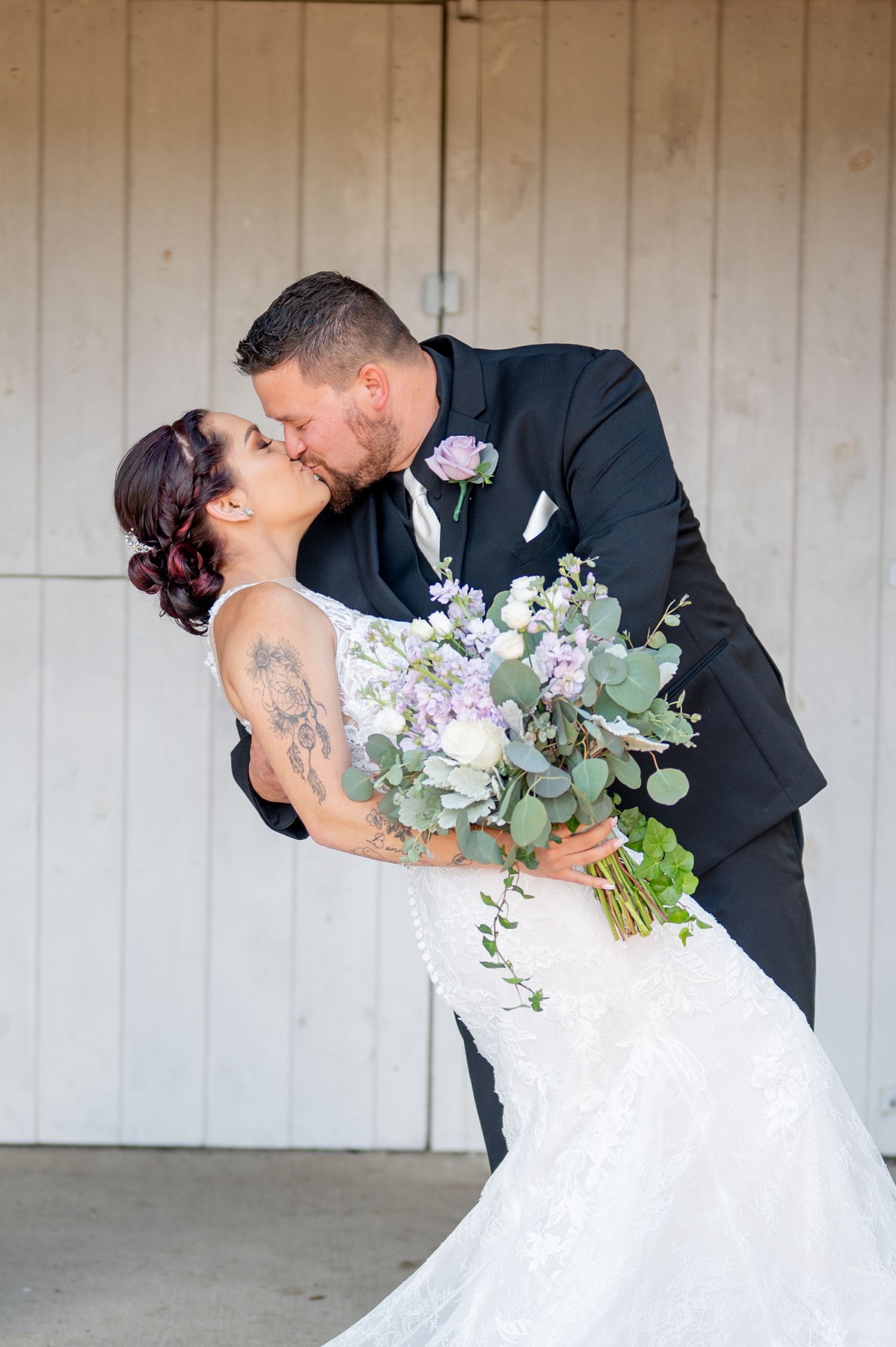 A romantic wedding photo of a couple sharing a kiss while holding a bouquet of eucalyptus and pale purple flowers.