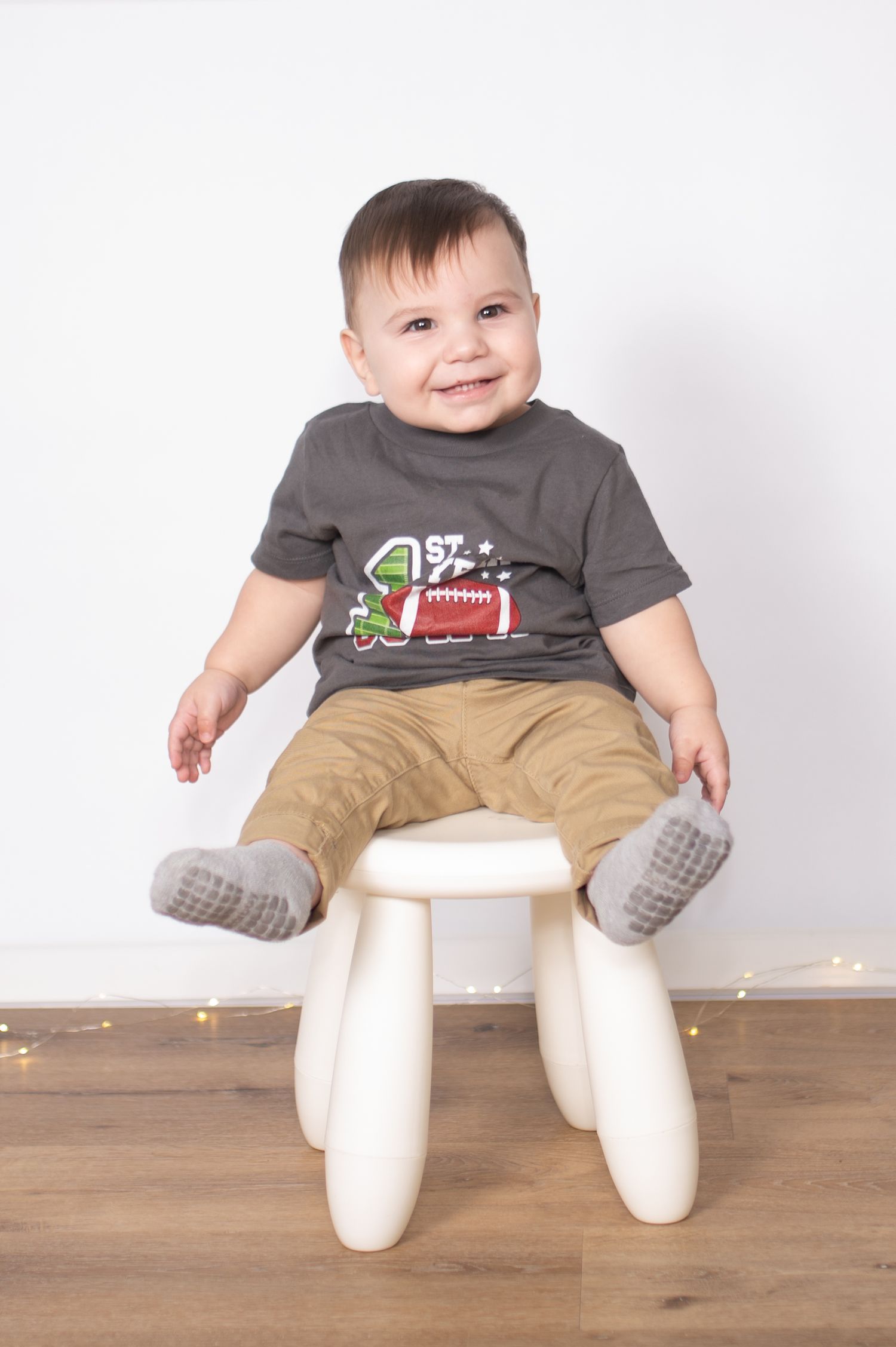 Baby in gray football t-shirt sits on wooden floor with legs spread out in playful pose against white background.