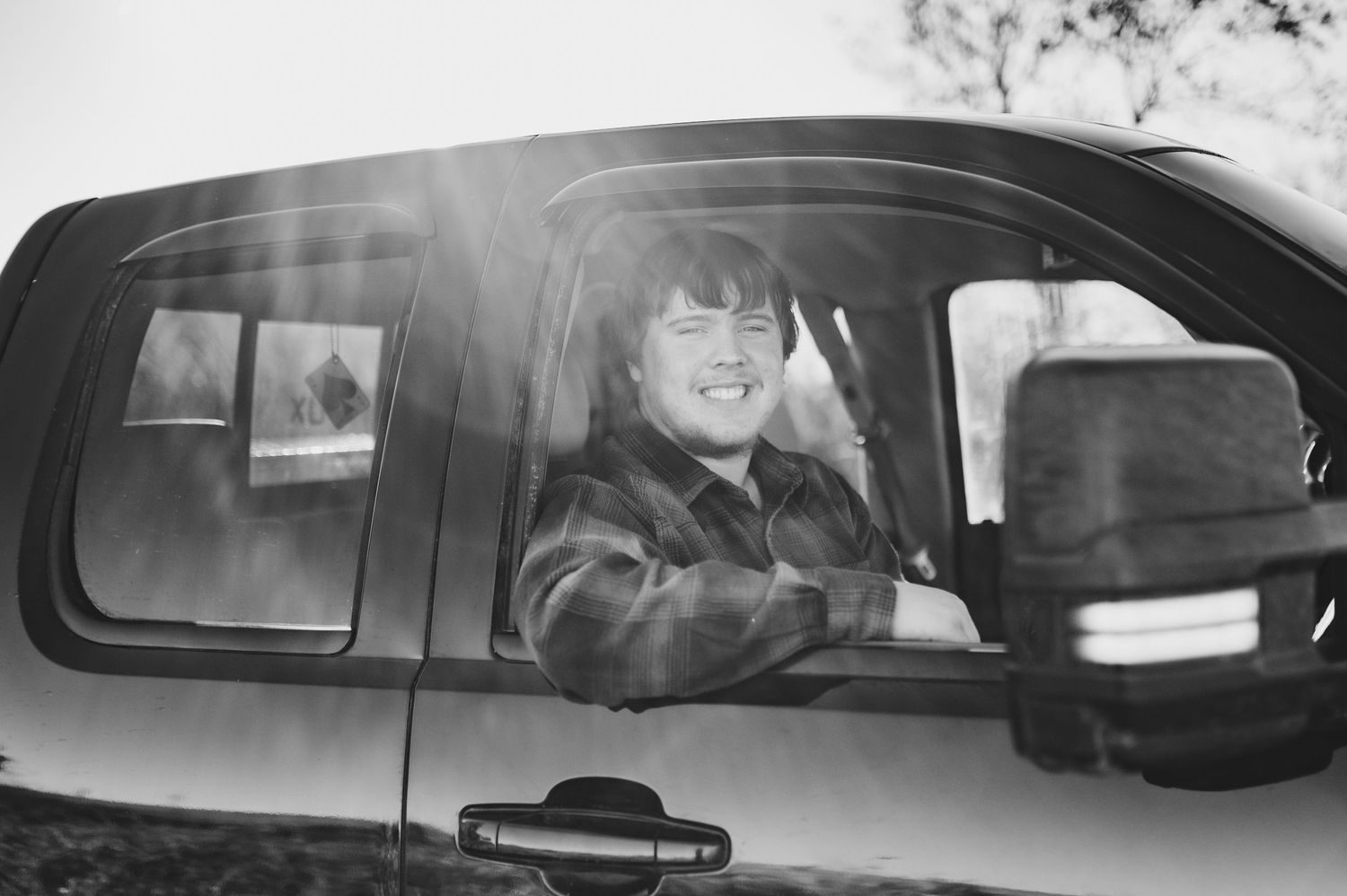 Black and white photo of someone smiling and waving while sitting in the driver's seat of a vehicle.