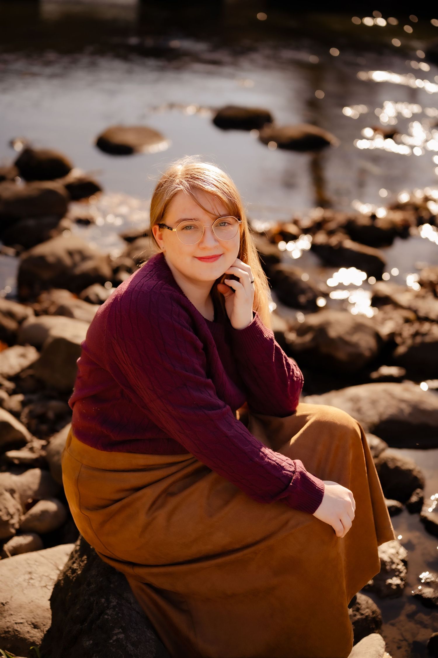 Person in a burgundy sweater sits thoughtfully on rocks by water with dramatic lighting and reflections.