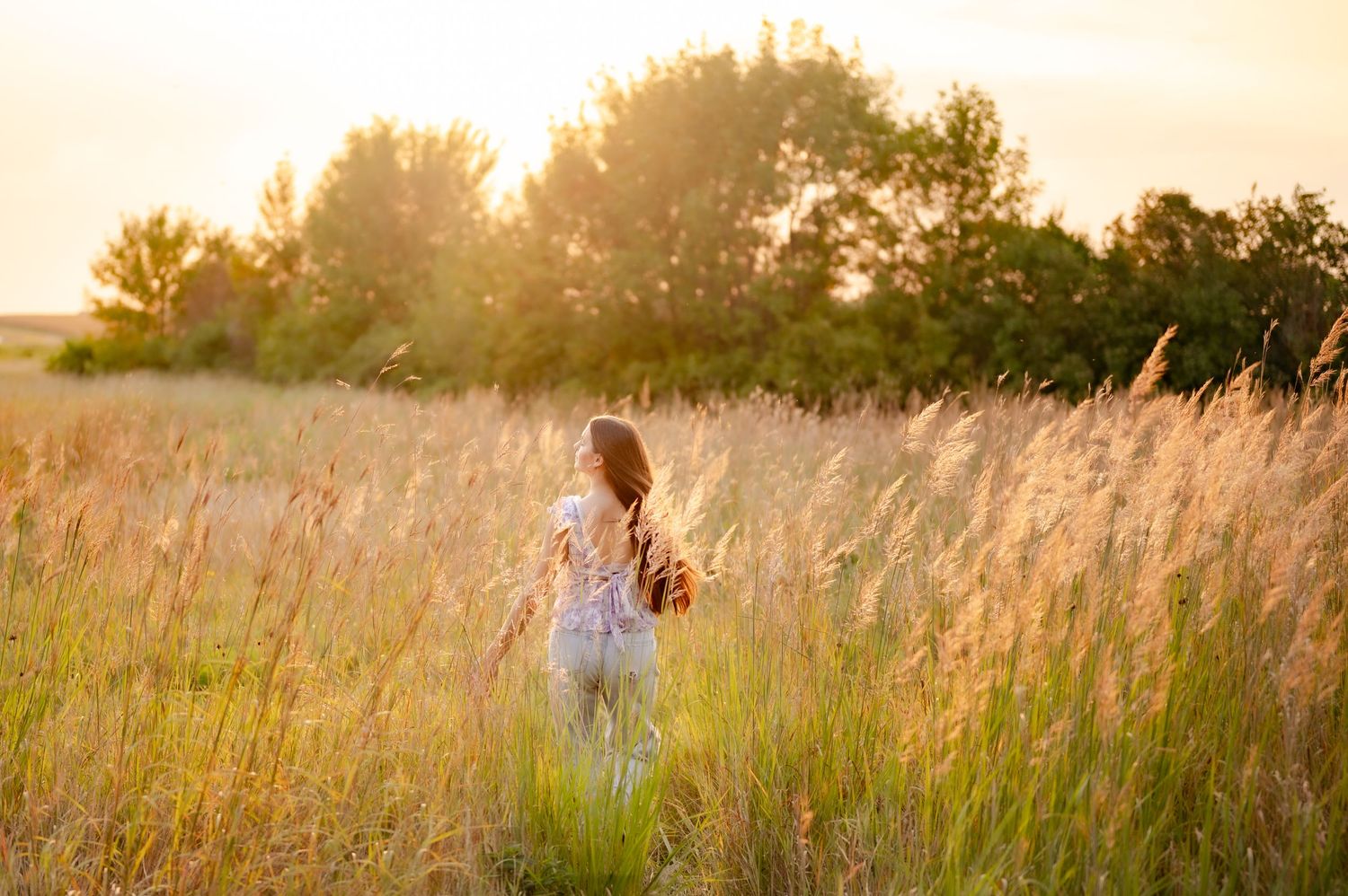 A silhouette stands among tall grass in a golden meadow at sunset with warm summer light filtering through.