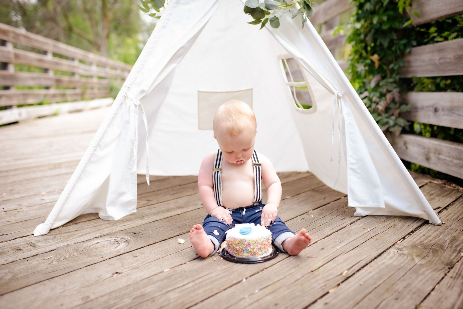 A white teepee tent set up on wooden deck with baby and cake inside.