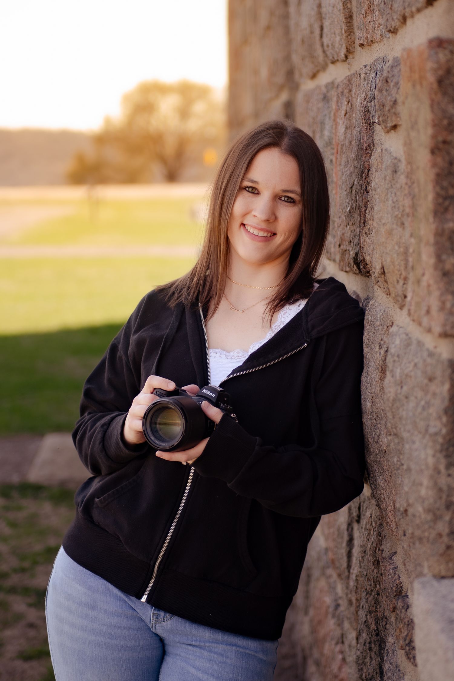 A photographer in a black hoodie and jeans holds a camera while standing against a stone wall at sunset.
