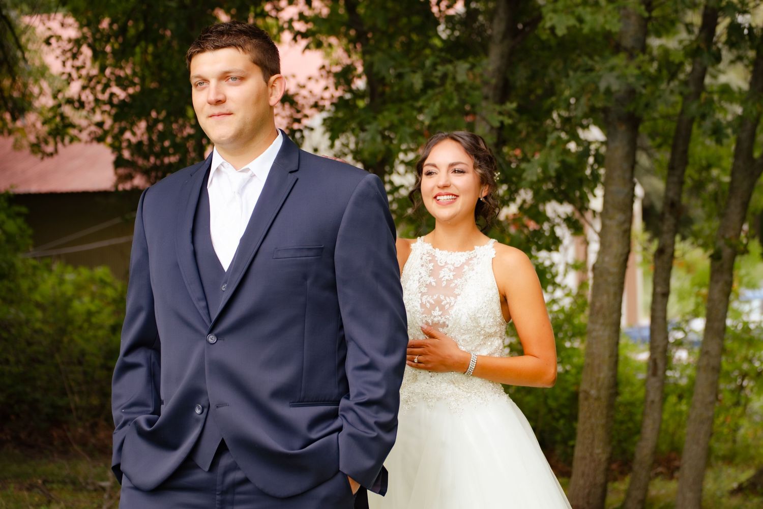 Wedding couple poses in wooded setting with bride in lace gown and groom in navy suit.