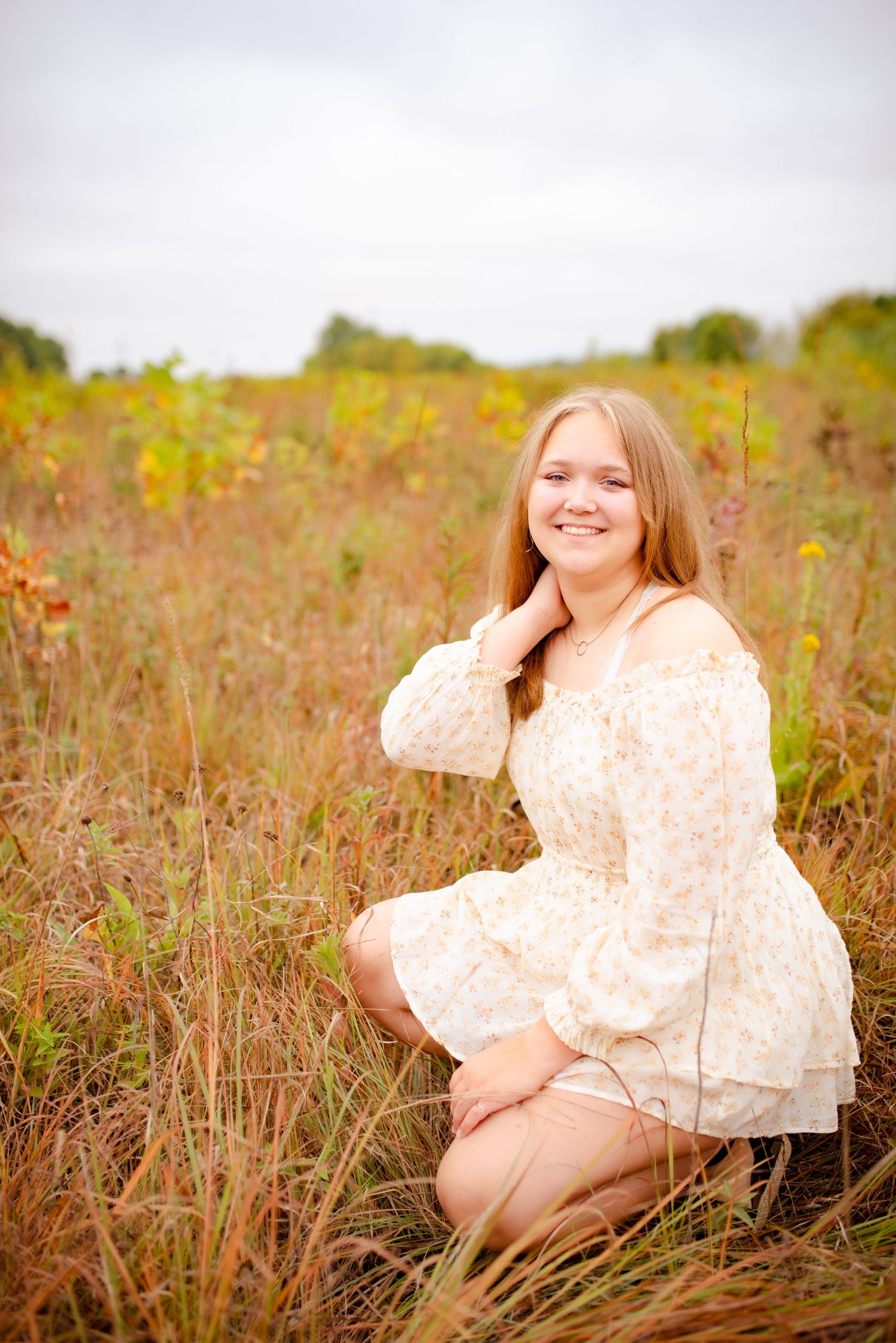 A person in a vintage-style cream dress kneels gracefully in a rustic field setting.