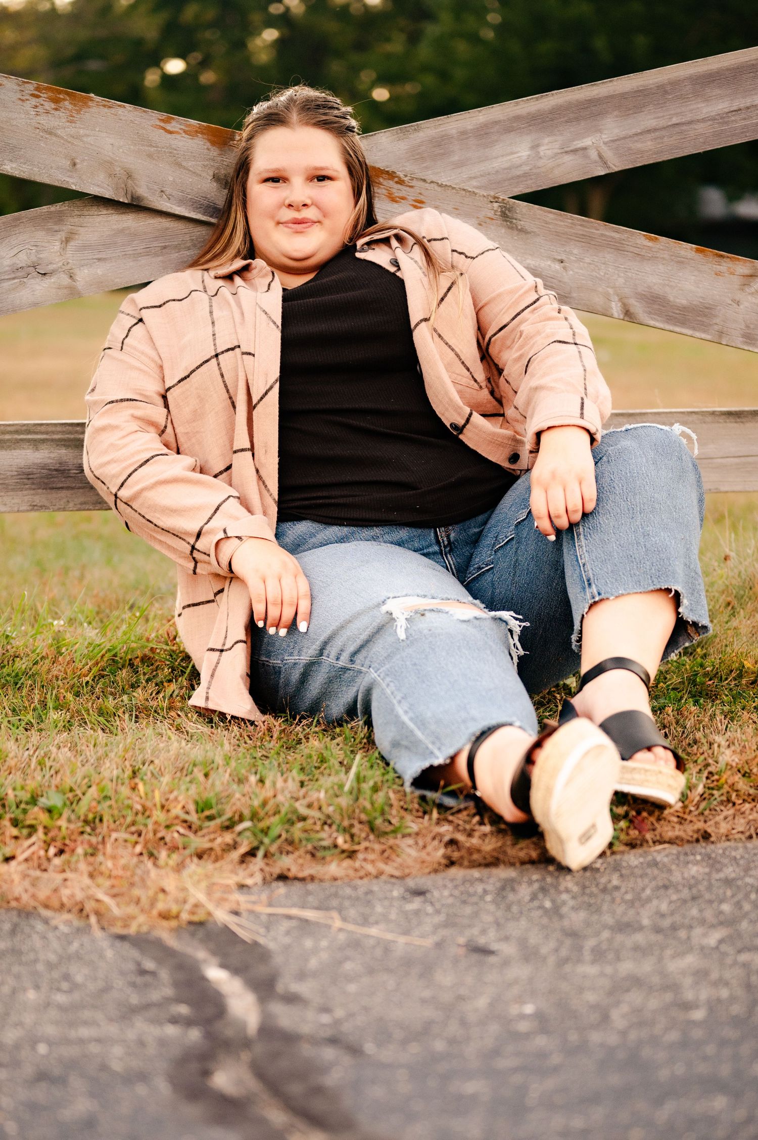 Person sitting casually on wooden fence wearing tan jacket, black top and distressed jeans.