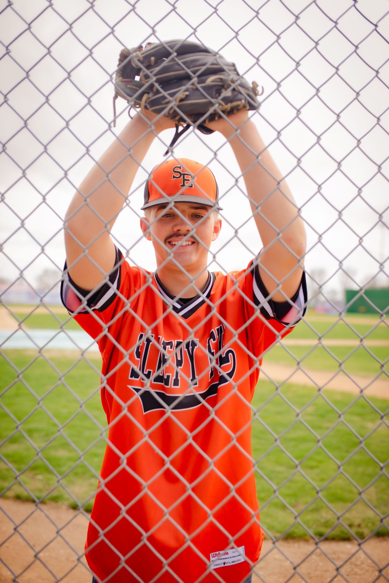 Baseball player in orange jersey poses behind chain link fence with glove raised overhead during practice.