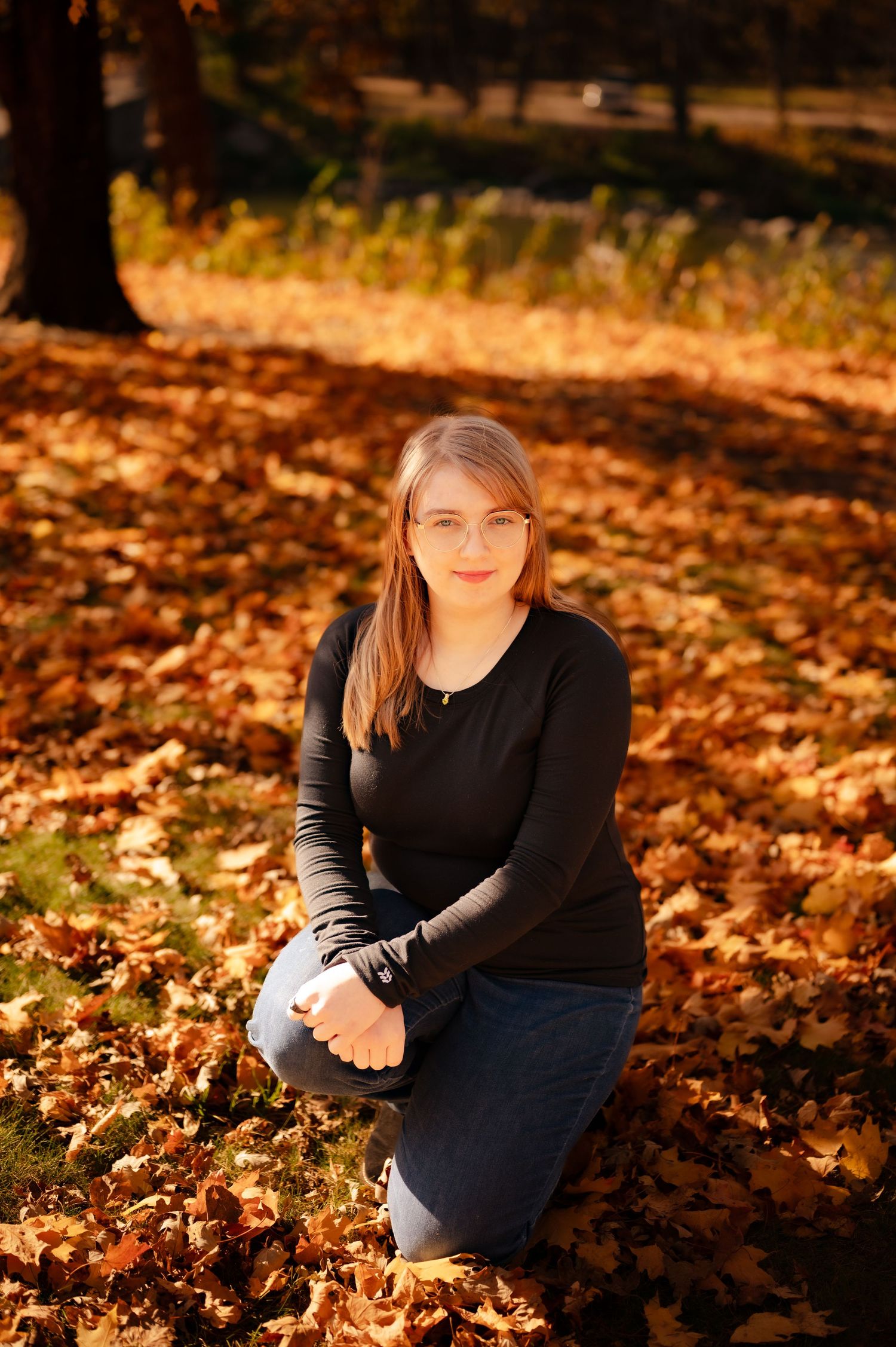 Portrait in autumn setting with fallen orange leaves on ground and warm sunlight filtering through trees.