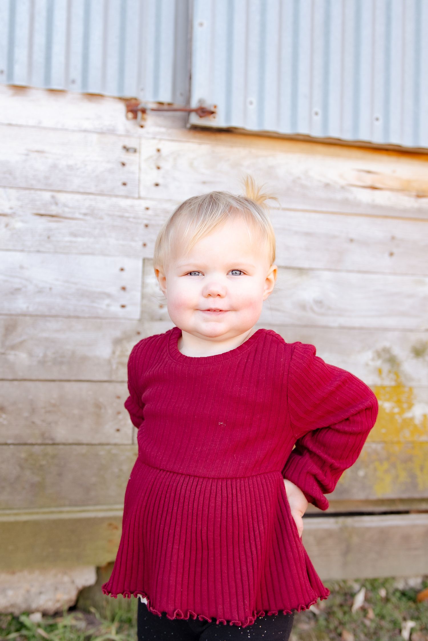 A small child in a red dress poses confidently against a rustic wall background.