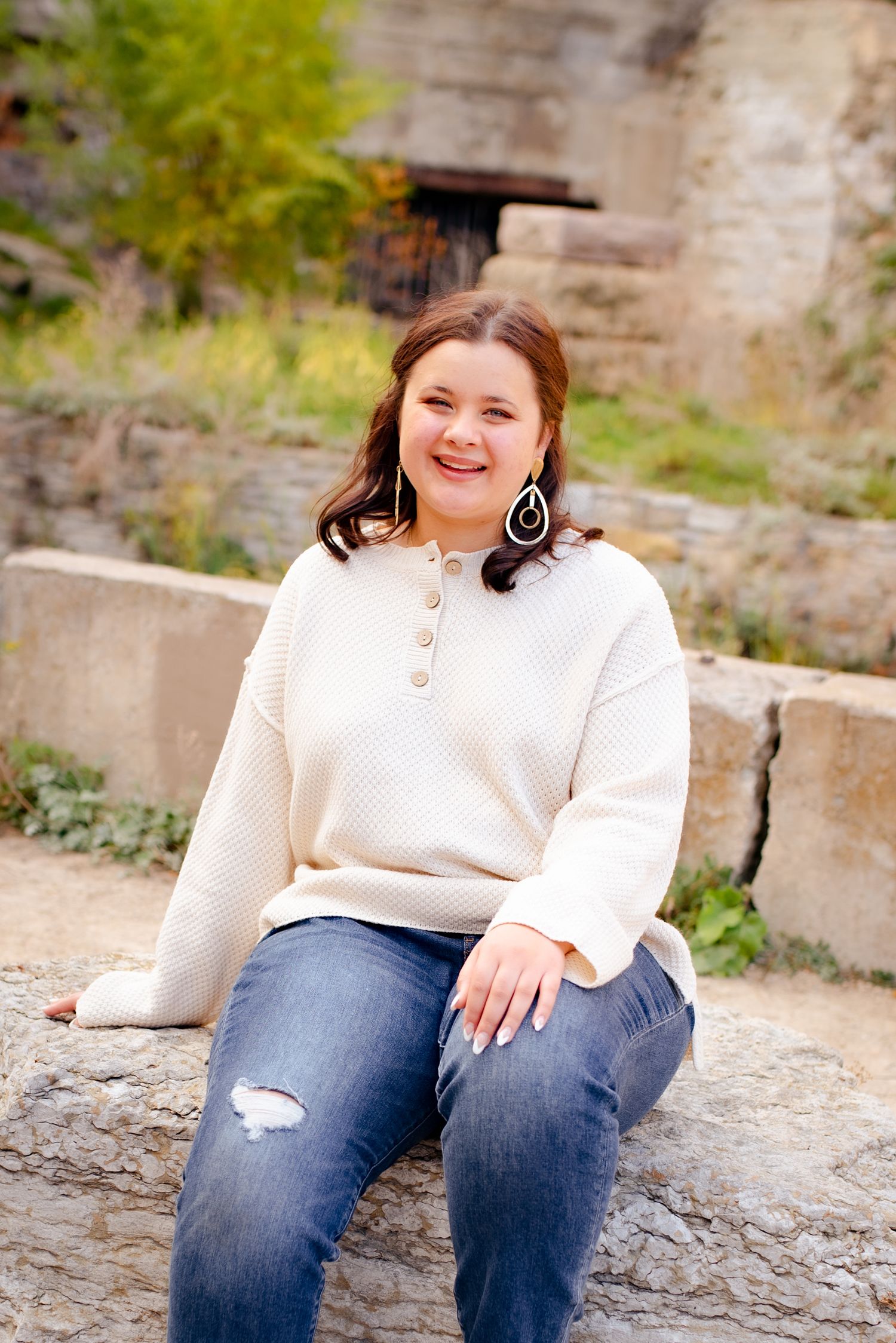 Person in white henley sweater sits casually on rock near stone bridge structure.