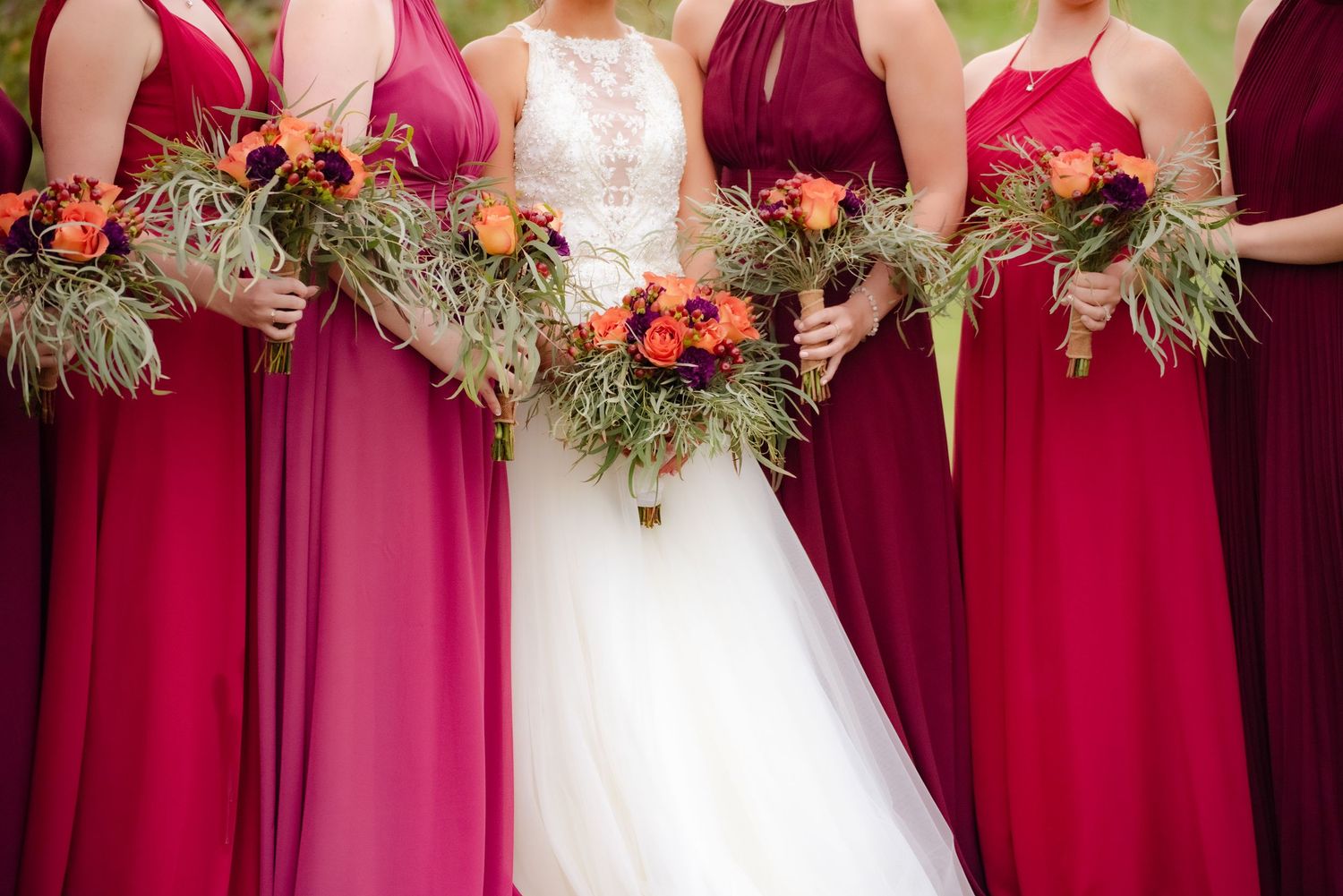 Bridesmaids in shades of burgundy and pink holding vibrant coral floral bouquets with greenery.