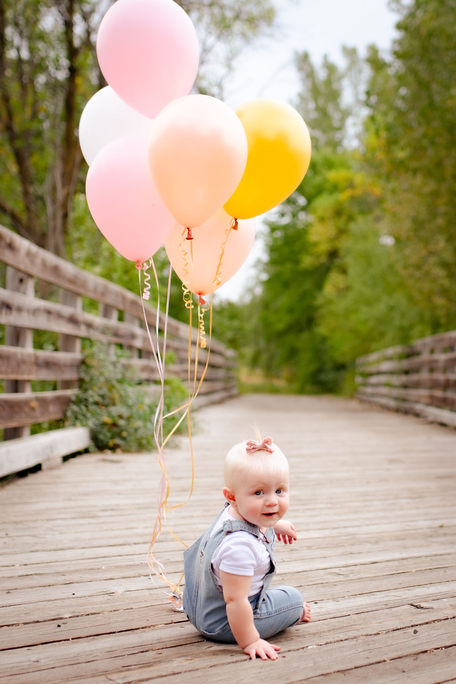 Baby in denim overalls sits on wooden boardwalk holding pink, peach and yellow balloons against nature backdrop.